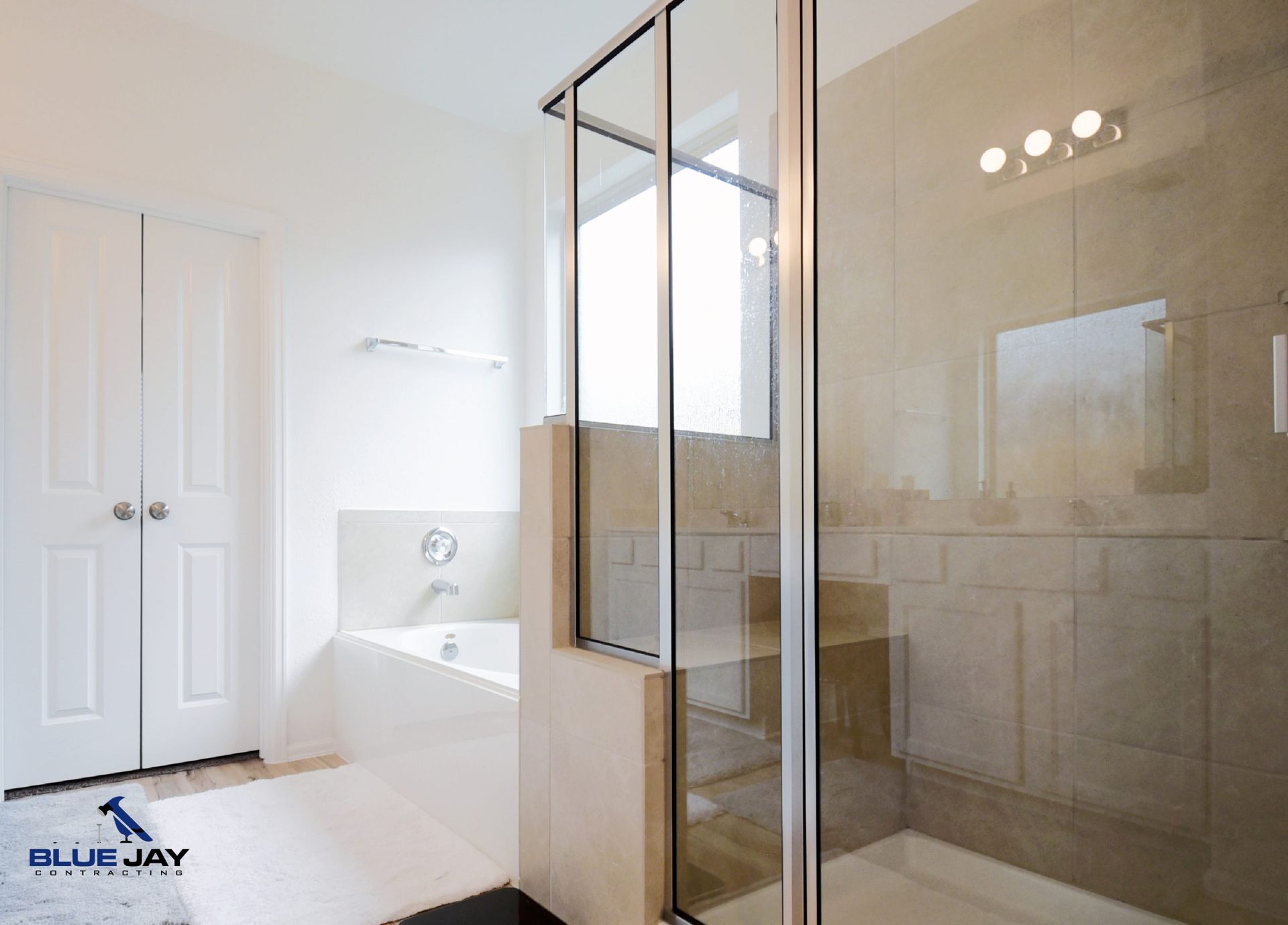 Bright bathroom with a glass shower, bathtub, and white doors. Soft beige tile and light fixtures.