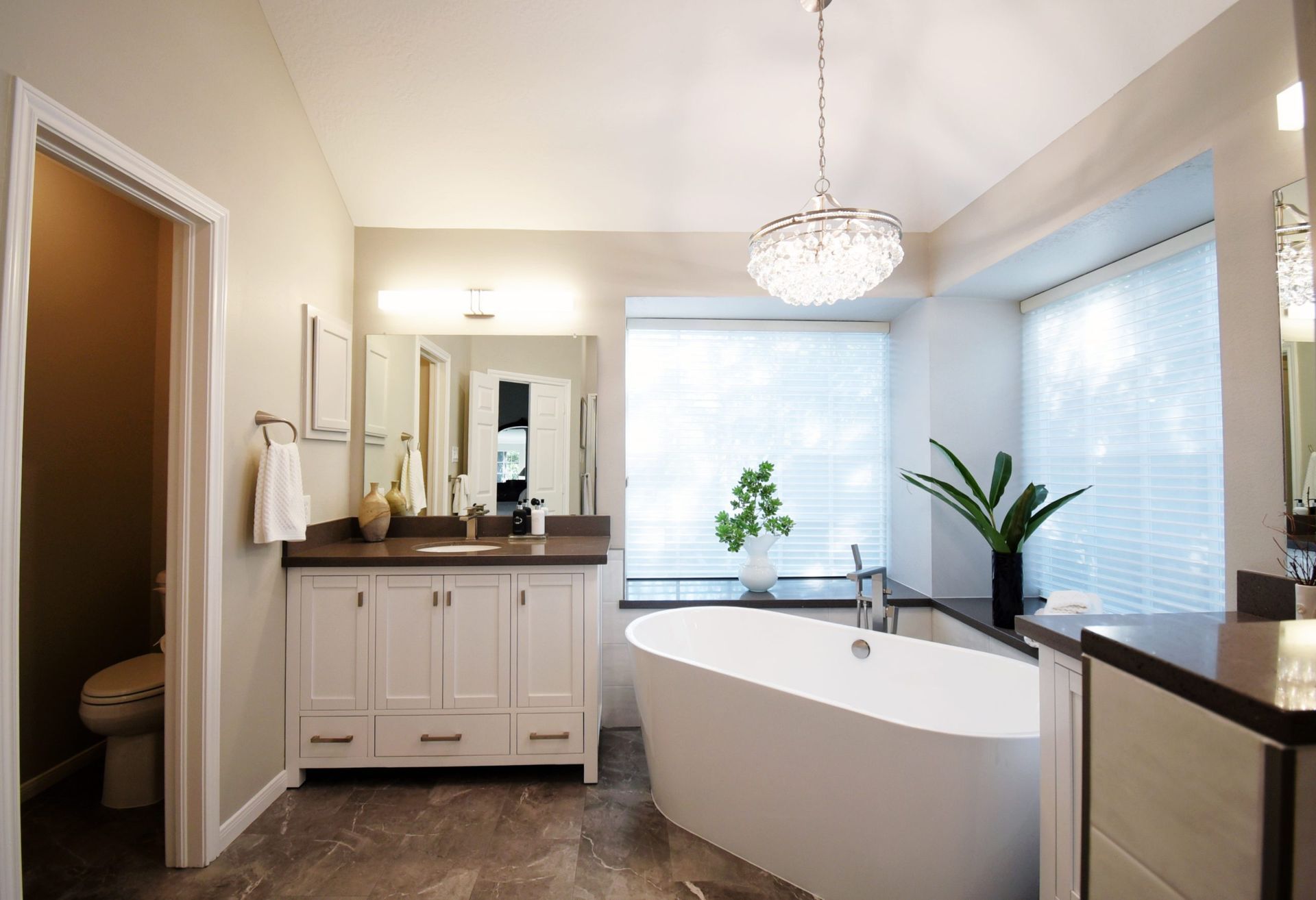 A modern bathroom with a white freestanding tub, white vanity, and large window.