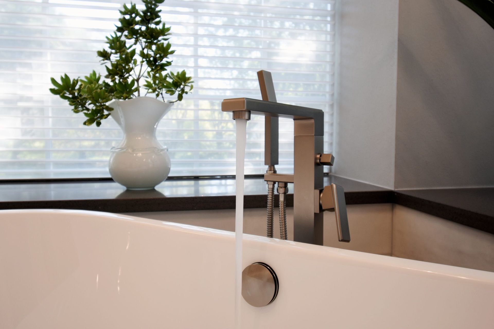 A bathtub with water flowing from a modern faucet; a white vase with greenery on a windowsill.