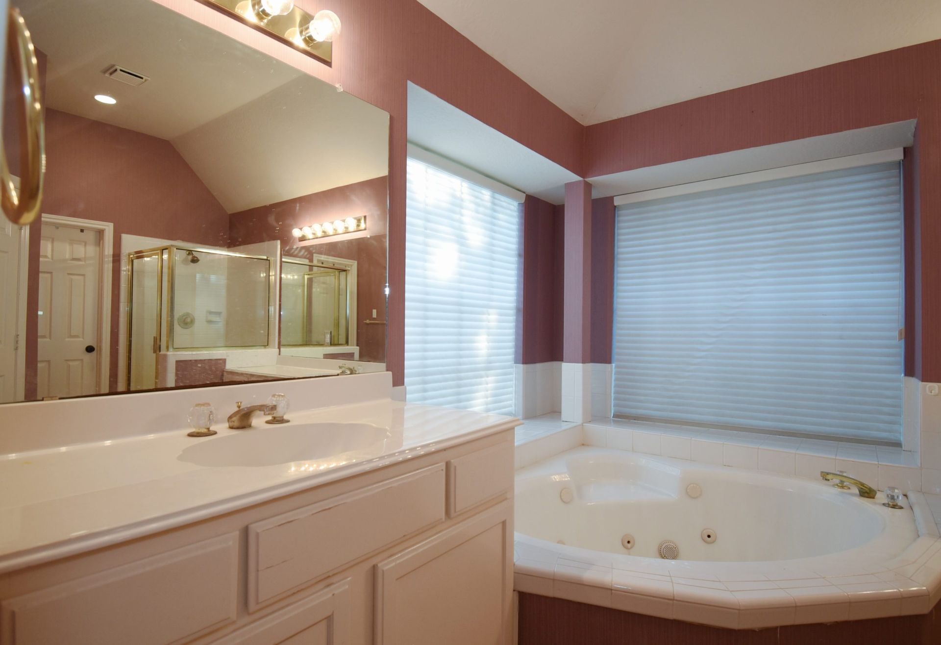 Bathroom with pink walls, white fixtures, large mirror, and a jacuzzi tub near a window with blinds.
