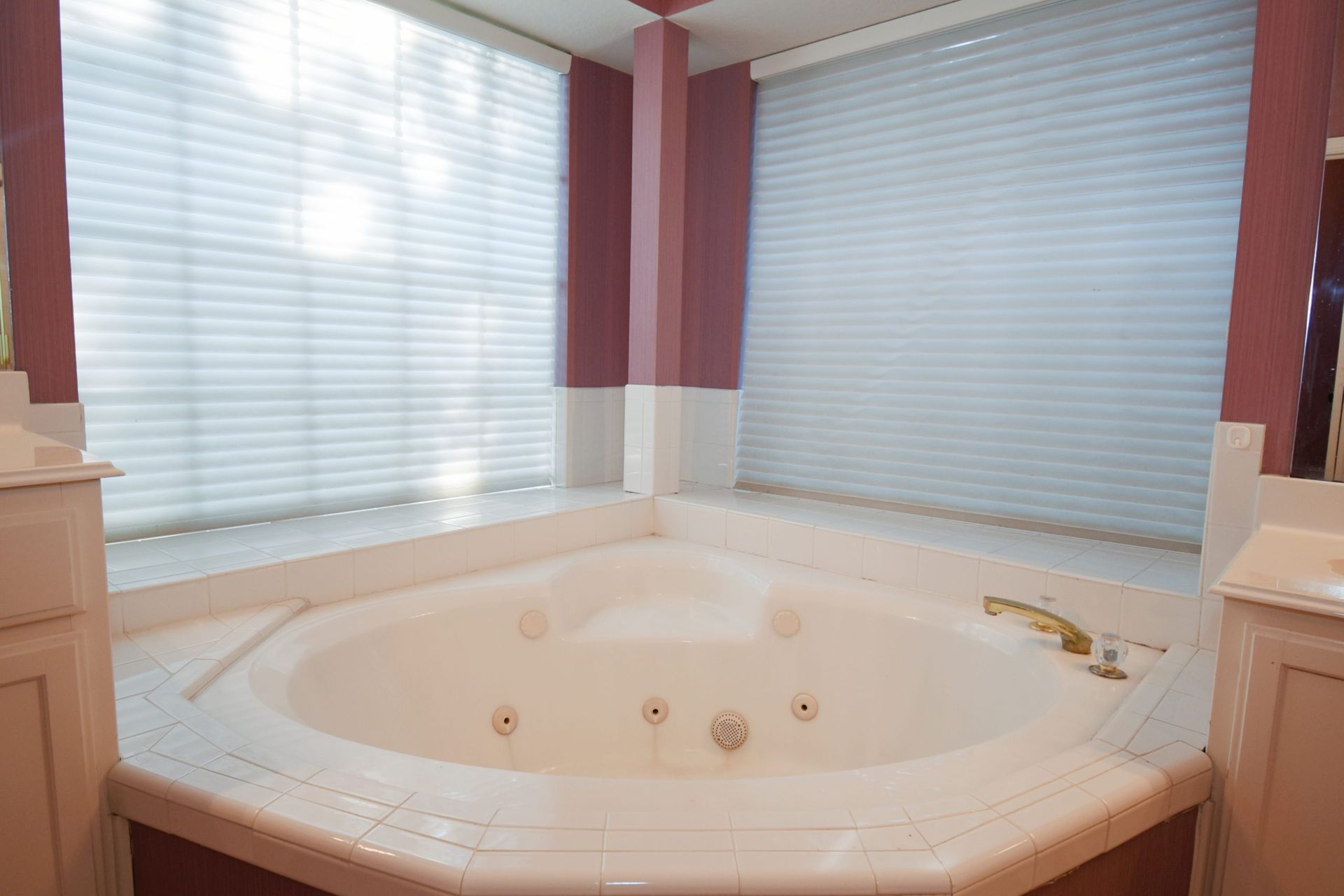 Triangular whirlpool tub in a bathroom corner, framed by two large windows with blinds.