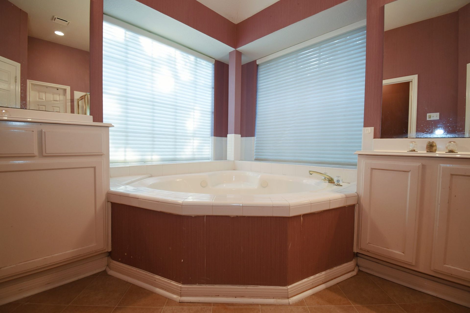 Bathroom with a jacuzzi tub in a corner, white cabinets, and large windows with blinds.