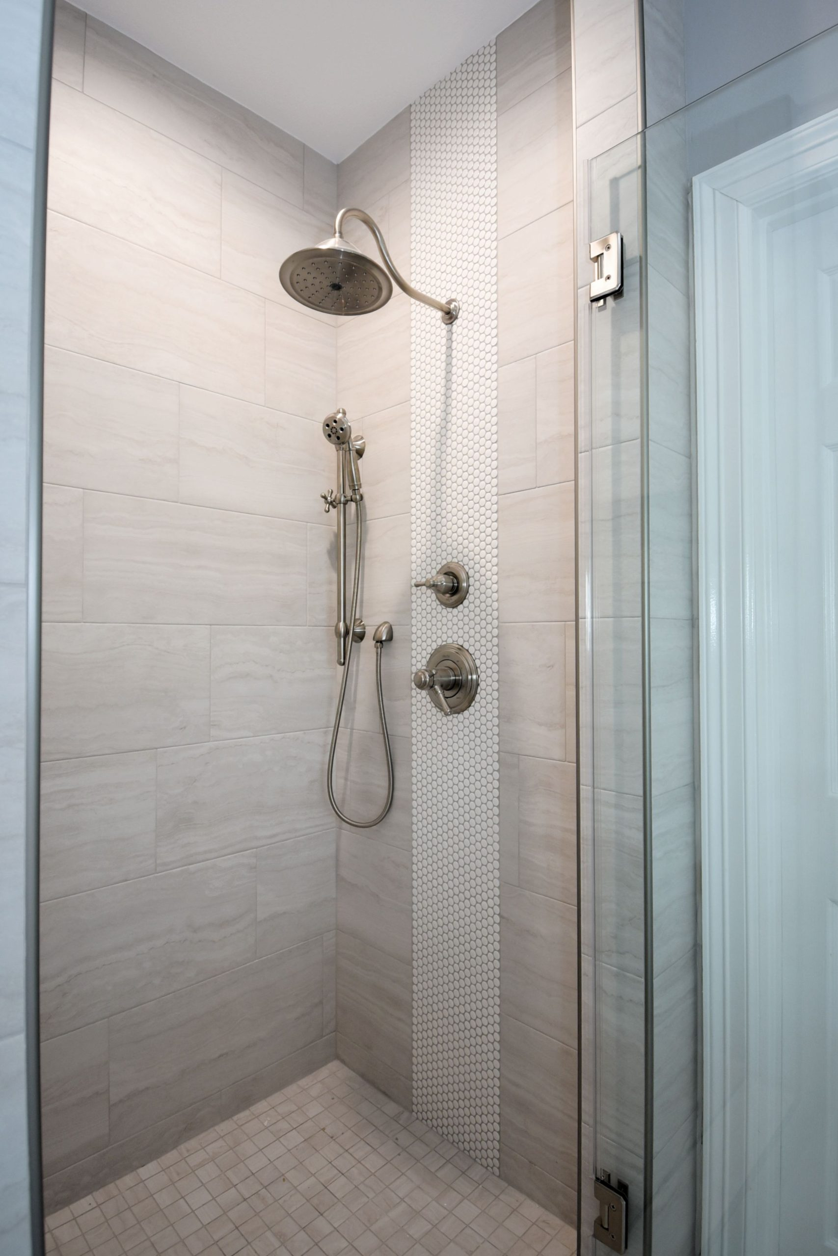 Shower stall with beige tile and glass door, featuring a rain showerhead and handheld sprayer.