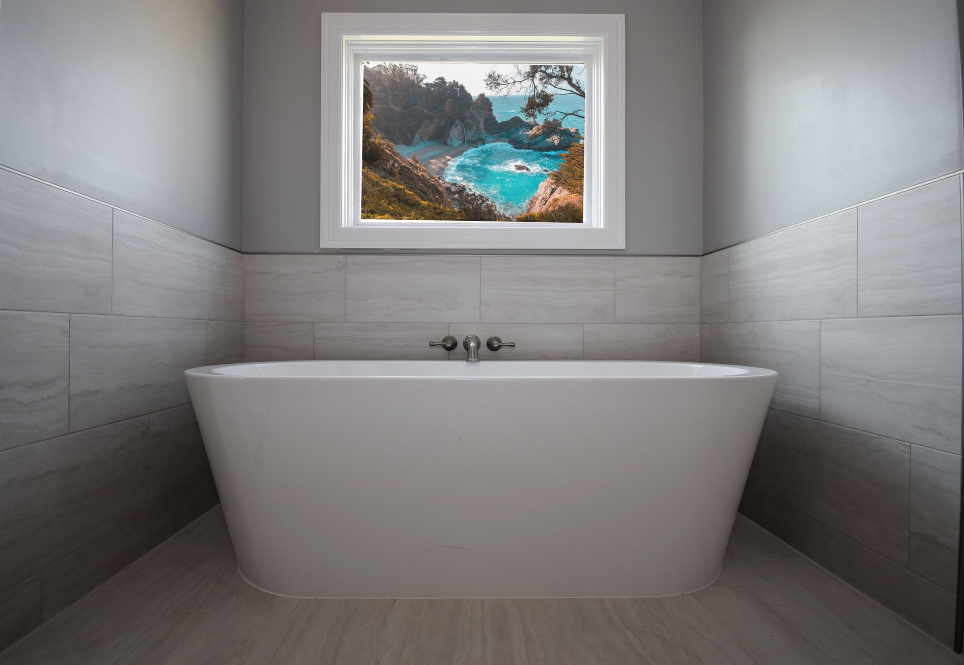 Freestanding white bathtub in bathroom with window overlooking a beach. Gray walls and light-colored floor.
