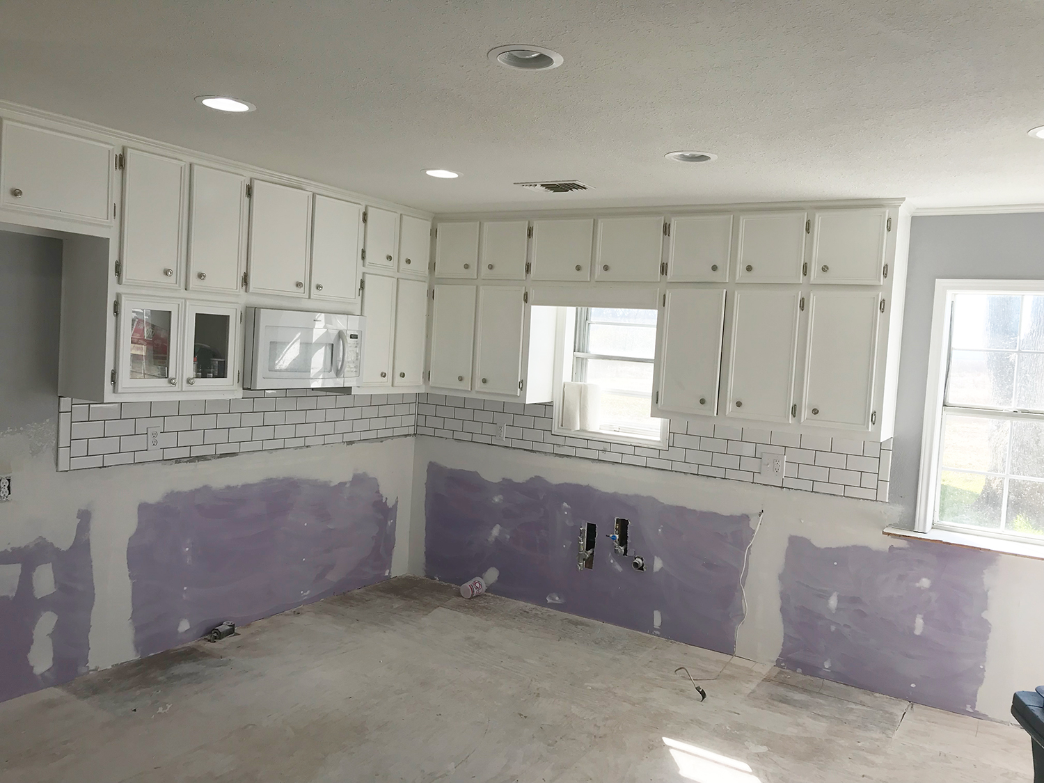 Kitchen under renovation: white cabinets, backsplash, exposed drywall, gray floor, and windows.