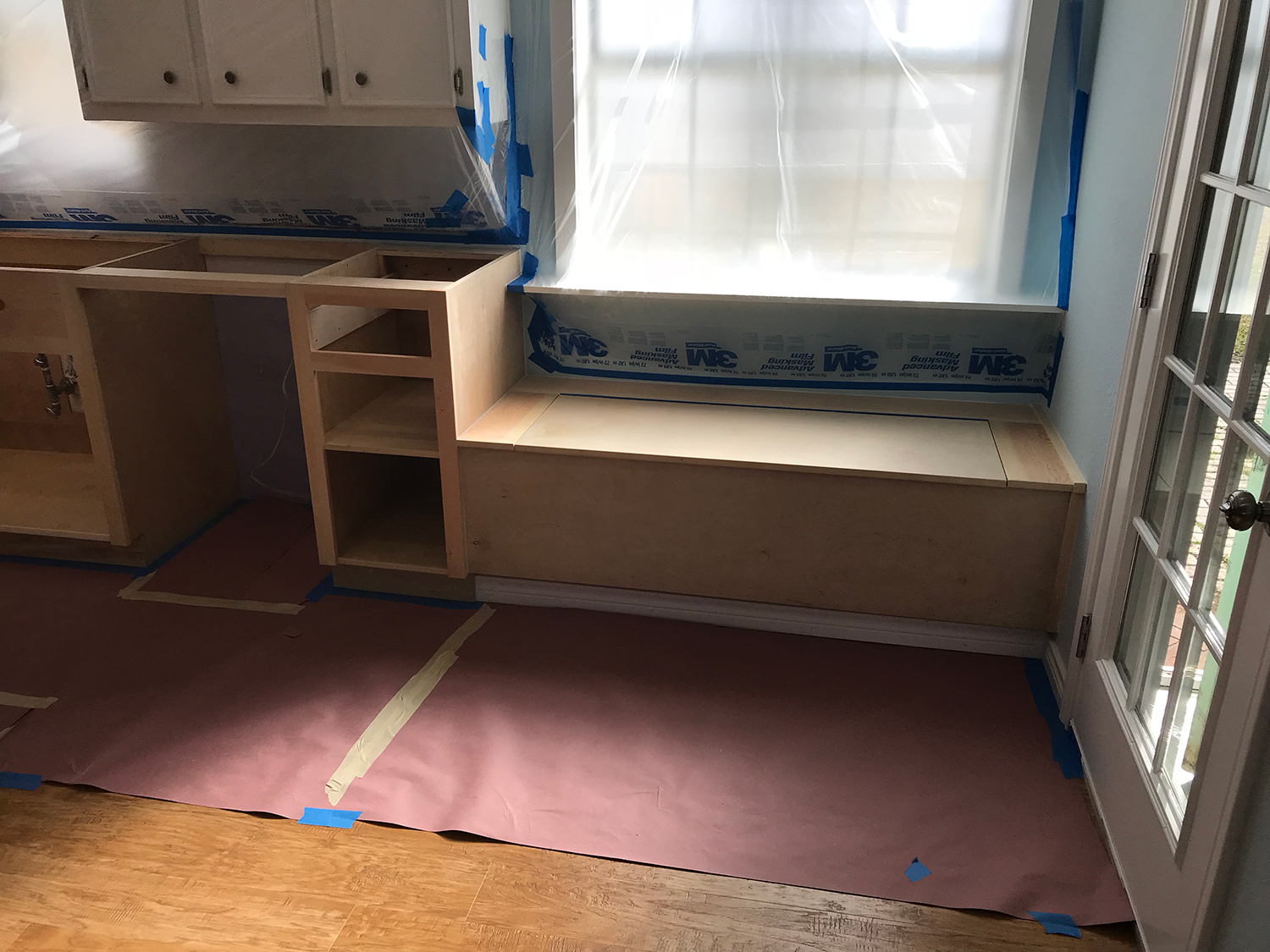 Kitchen with built-in bench and unfinished cabinets under construction, floor covered in protective paper, blue tape.