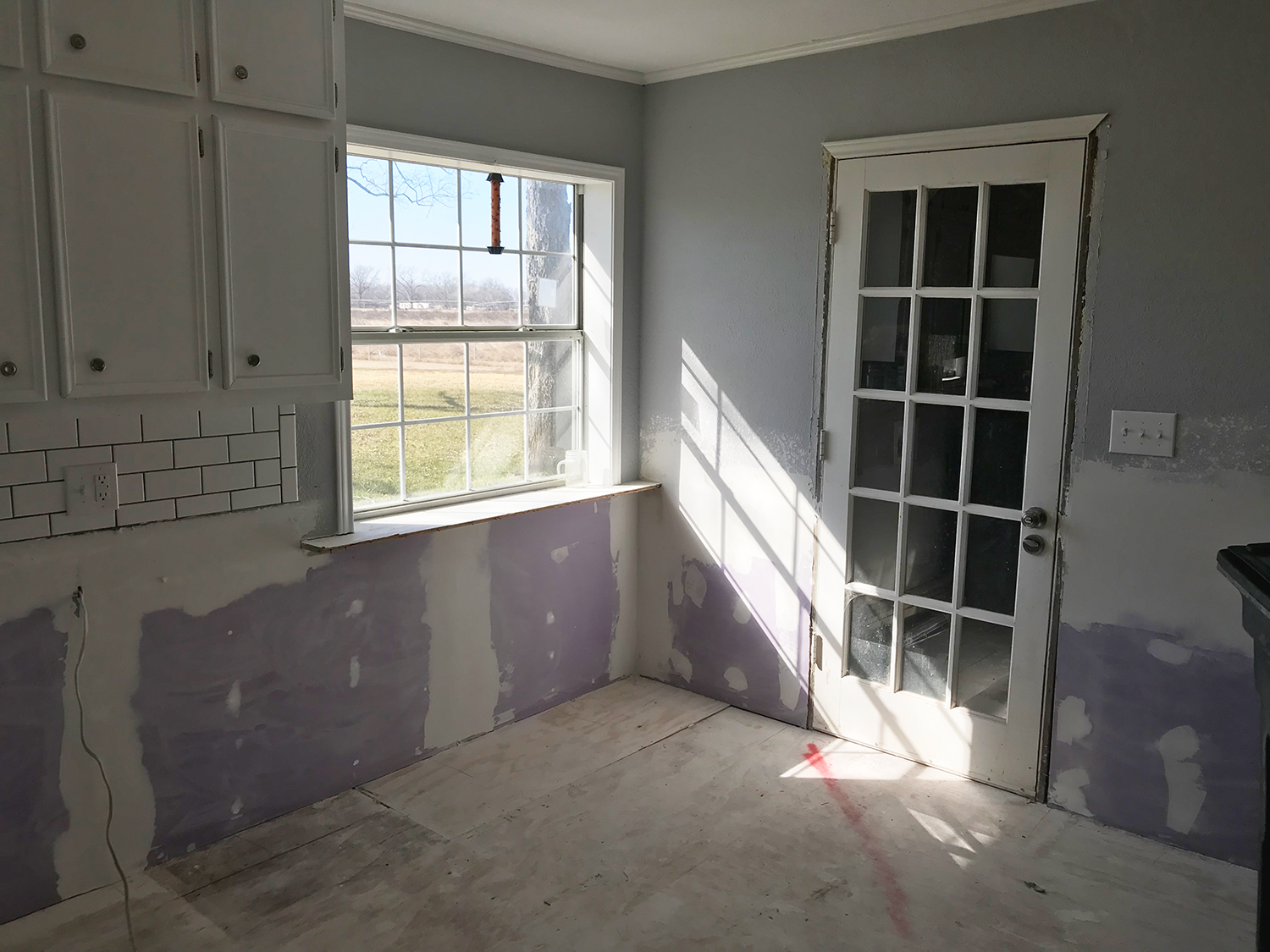 Kitchen with unfinished drywall, white cabinets, a window, and a glass-paned door.