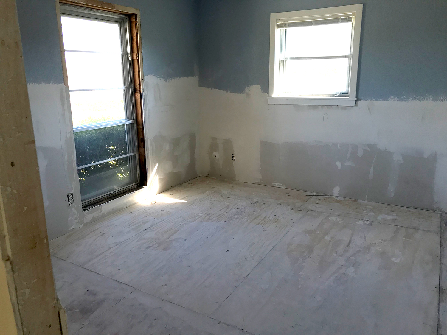 Room with partially painted blue walls and unfinished drywall. Sunlight streams through door.