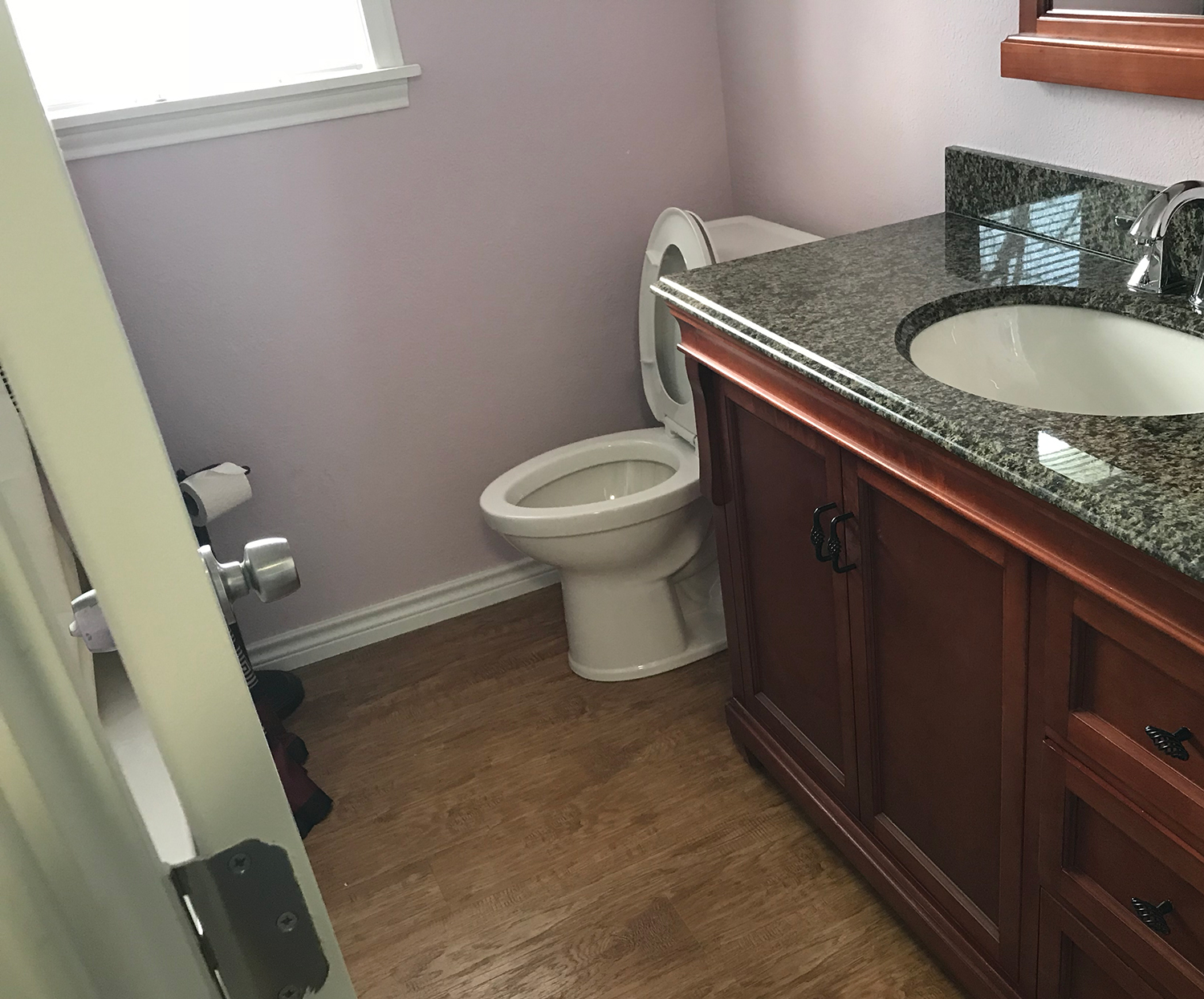 Bathroom with toilet, vanity, sink, and window; light purple walls, wood floor.