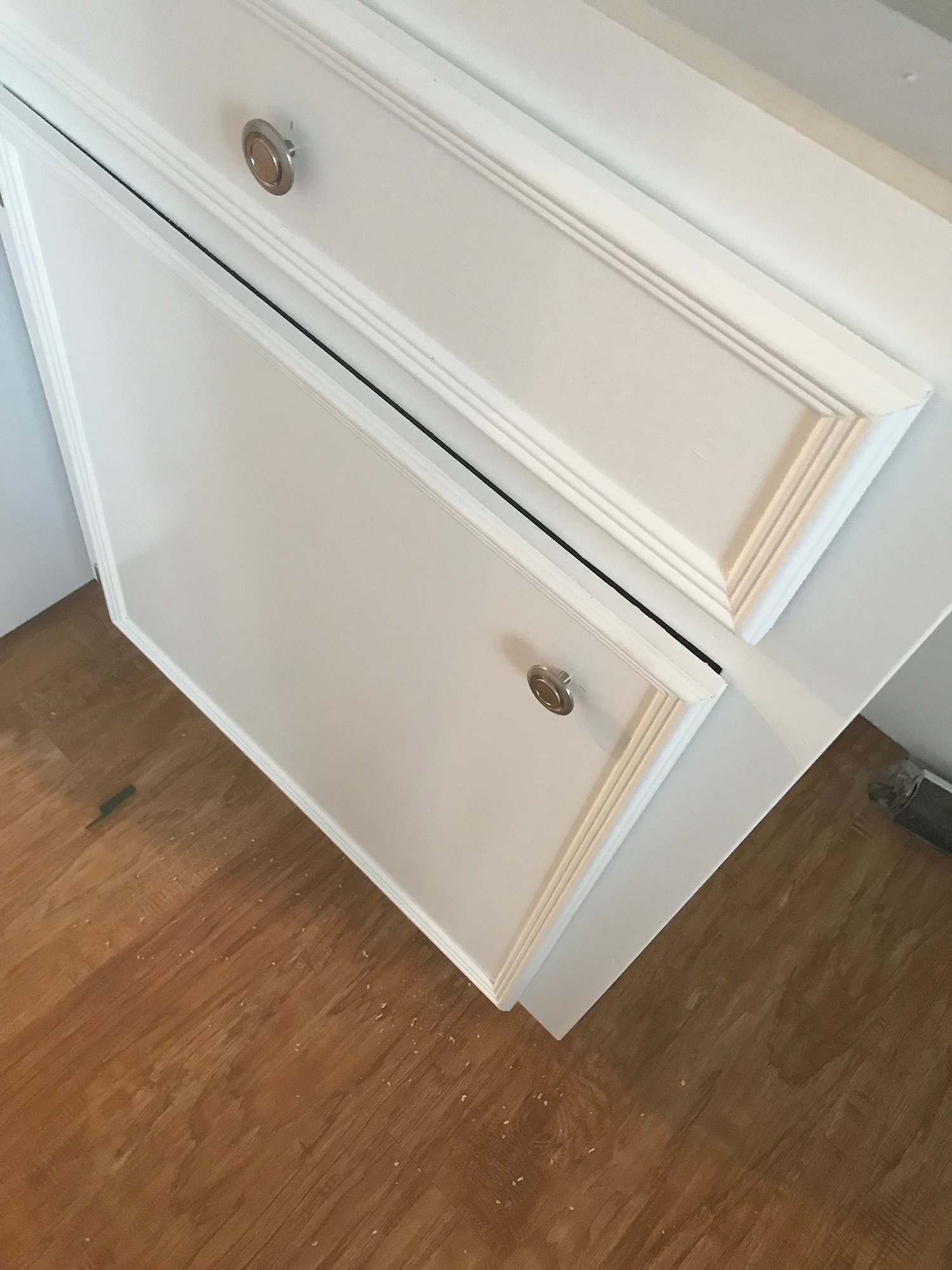 White kitchen cabinet with a drawer and door, silver knobs, wood floor.
