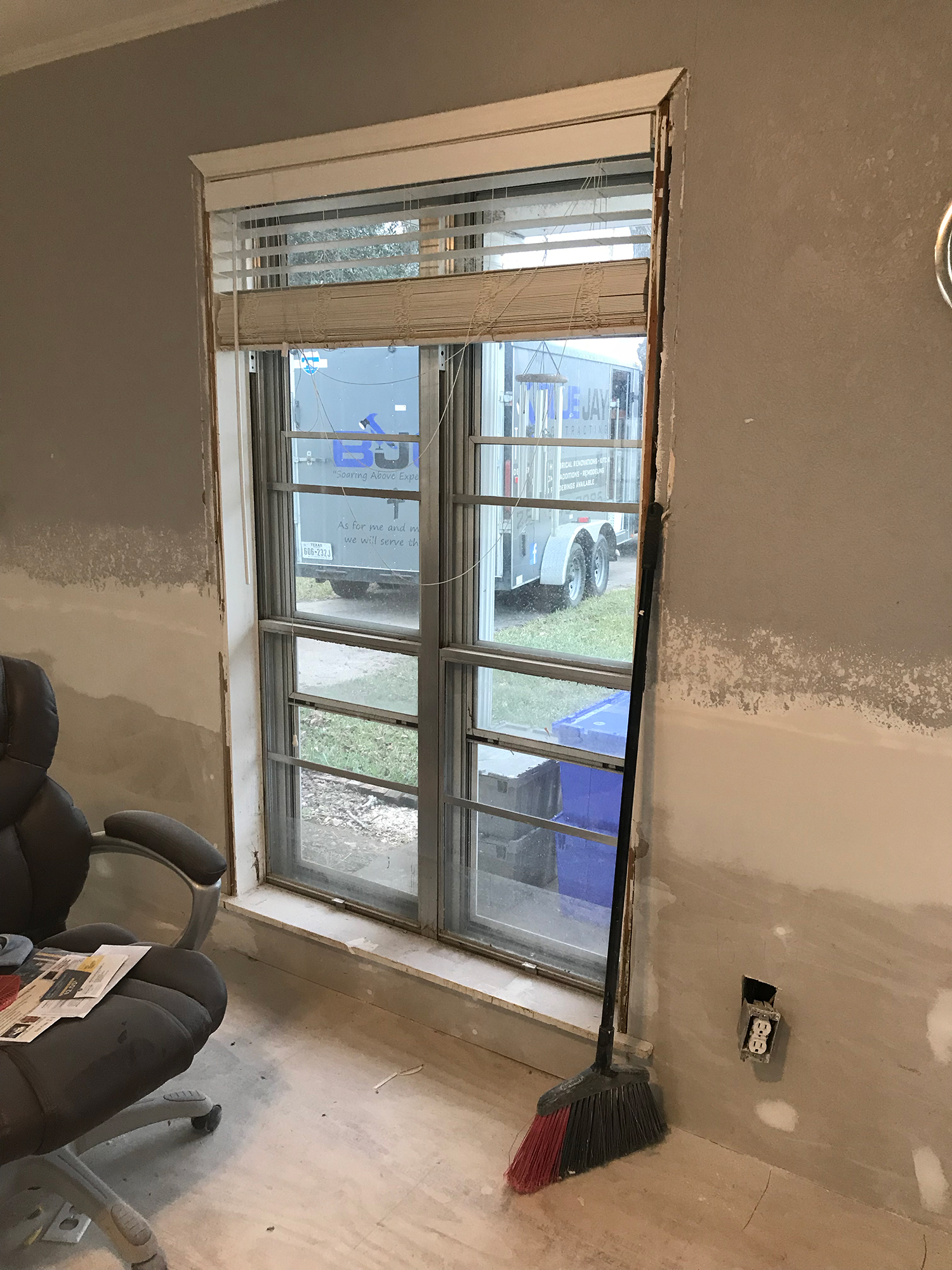 Window with blinds, in a room under construction. A broom leans against the frame.