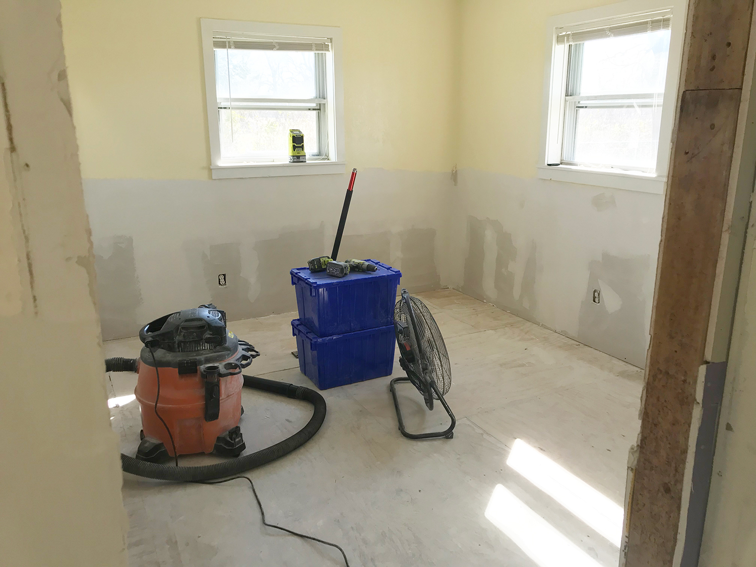 Room under renovation; drywall, vacuum, blue bins, fan, and two windows.