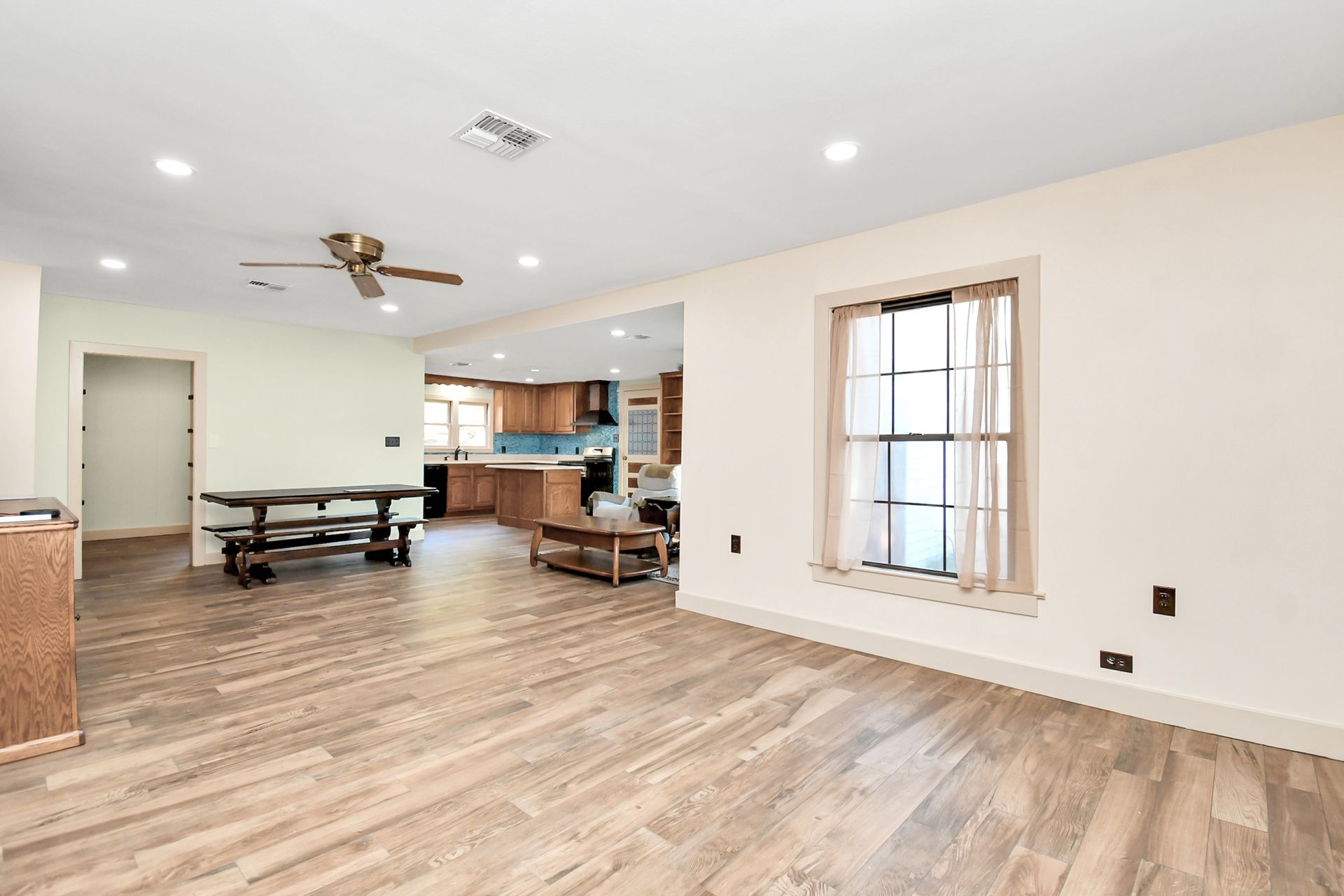 Open-plan living space with wood-look flooring, kitchen in the background, and a window with curtains.
