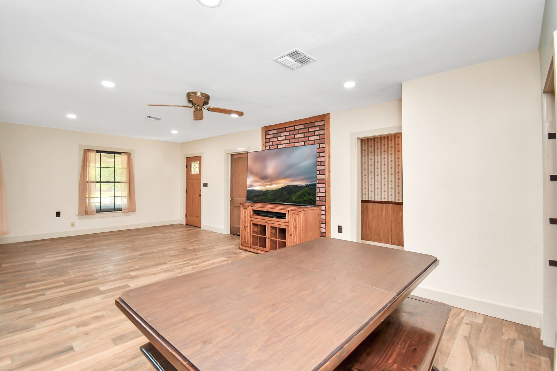 Open-plan living room with TV on wood stand, wooden floors, and a large table in the foreground.