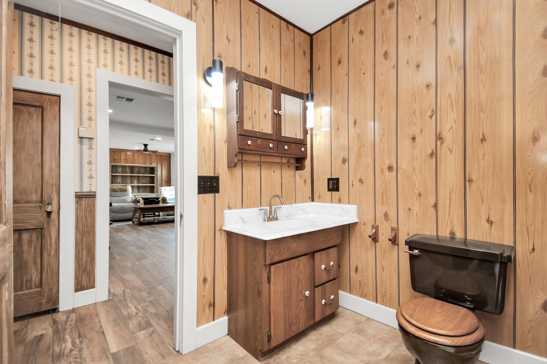 Bathroom with wood paneling, vanity, and toilet. View into a hallway with patterned wallpaper.