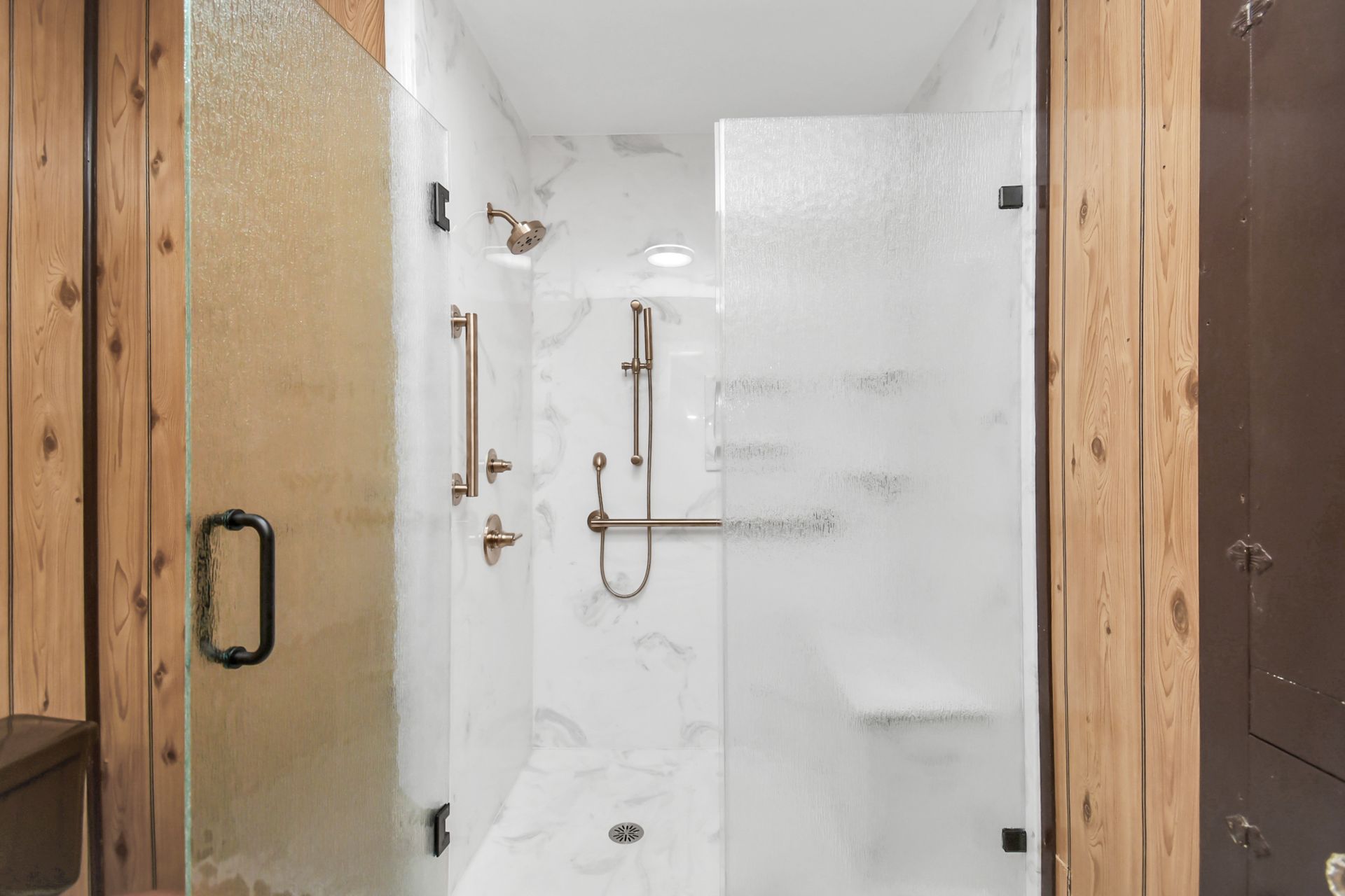 Accessible shower with frosted glass doors, marble-like tile, and grab bars in a wood-paneled room.
