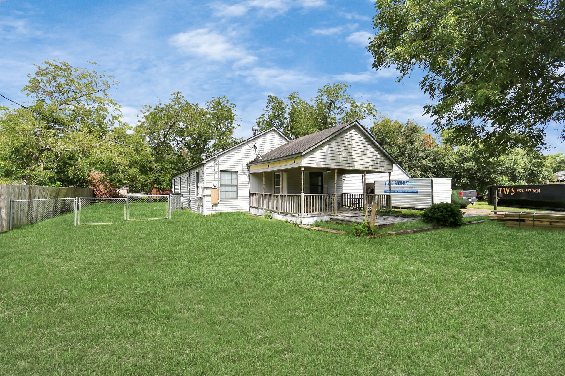 A small, weathered house with a porch in a grassy yard, surrounded by trees and a fence.