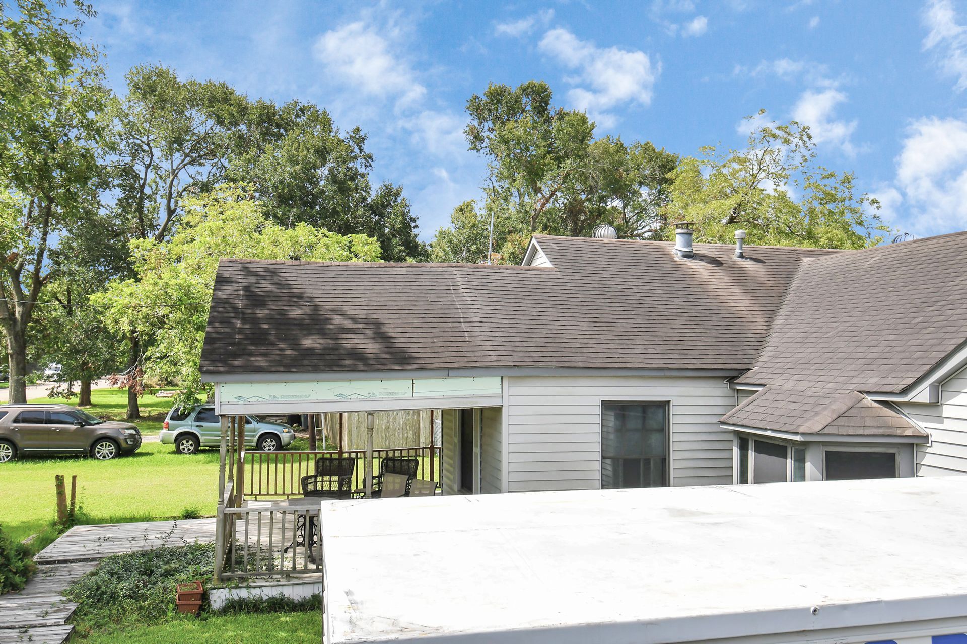 A weathered house with a covered porch and cars parked on the green lawn on a sunny day.