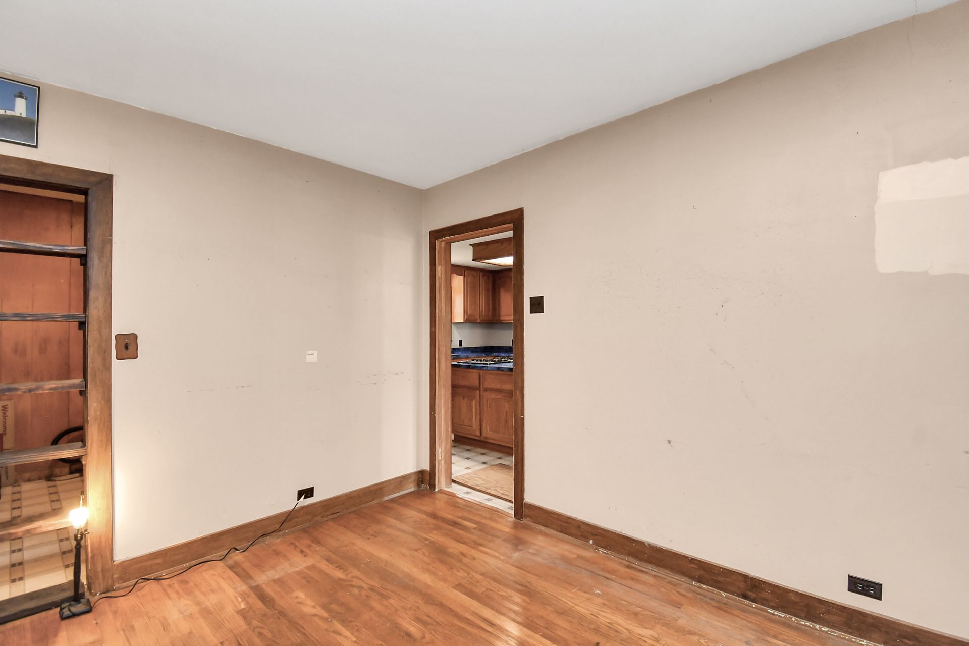 Empty room with hardwood floors, doorway to kitchen, built-in bookshelves, and beige walls.