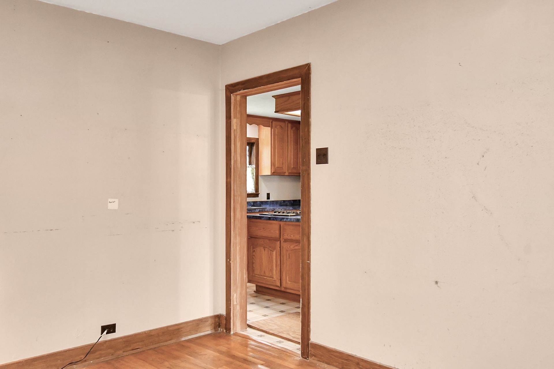 An empty room with wooden floors and an open doorway leading to a kitchen with wooden cabinets.
