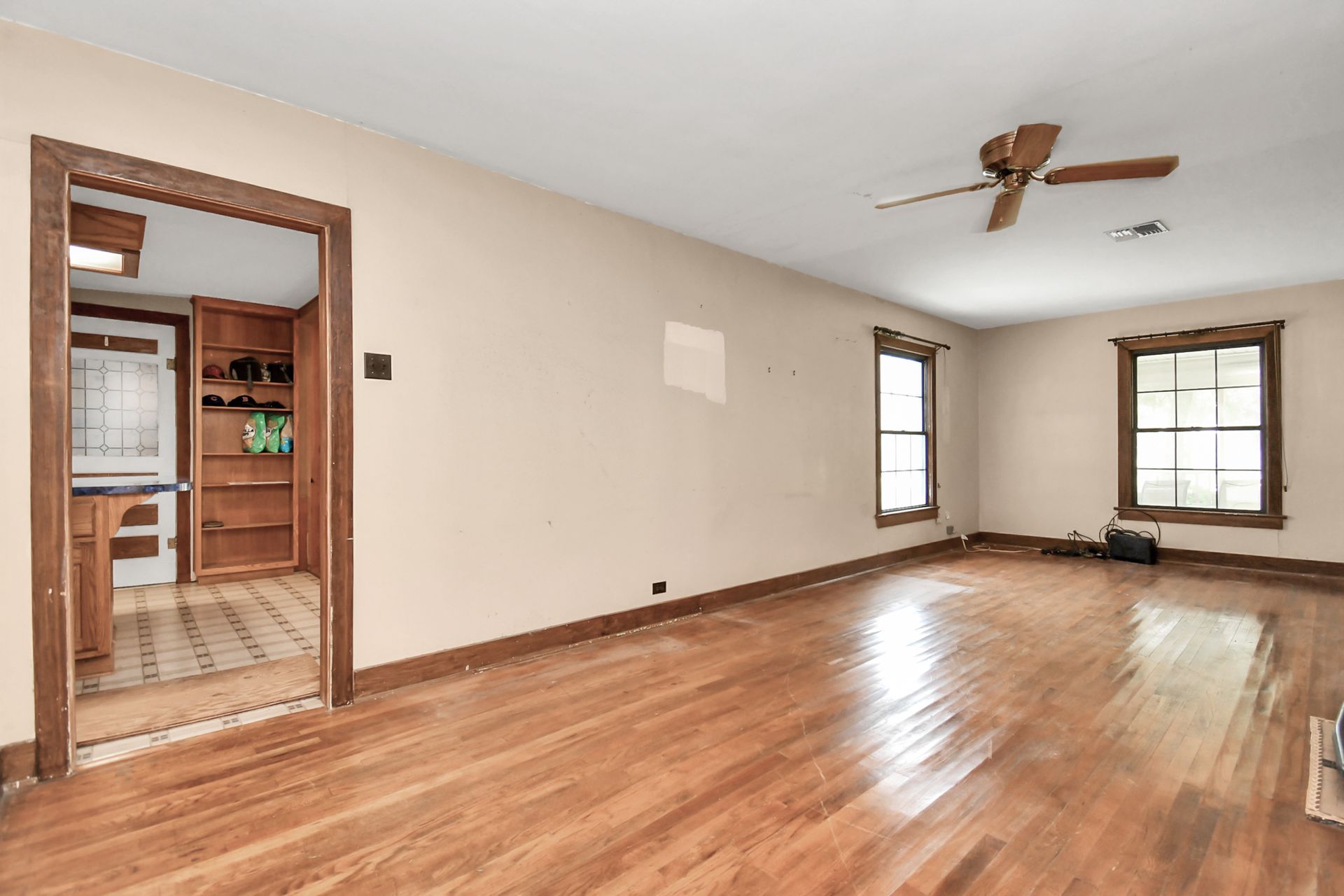 Empty living room with hardwood floors, beige walls, and a doorway leading to a kitchen.