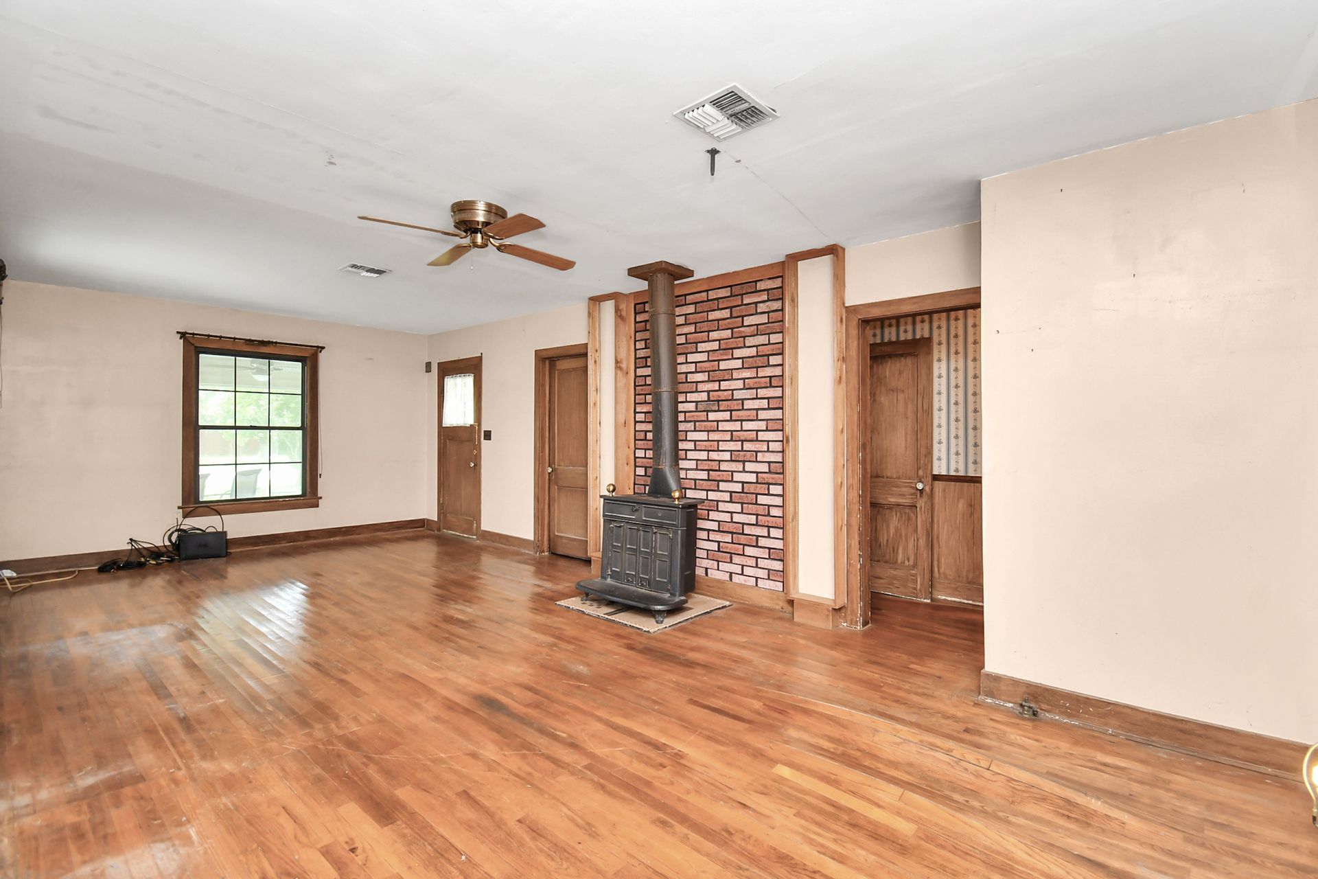 Empty room with wood floors, brick chimney, and wood-burning stove. Beige walls, wood doors, and a ceiling fan.