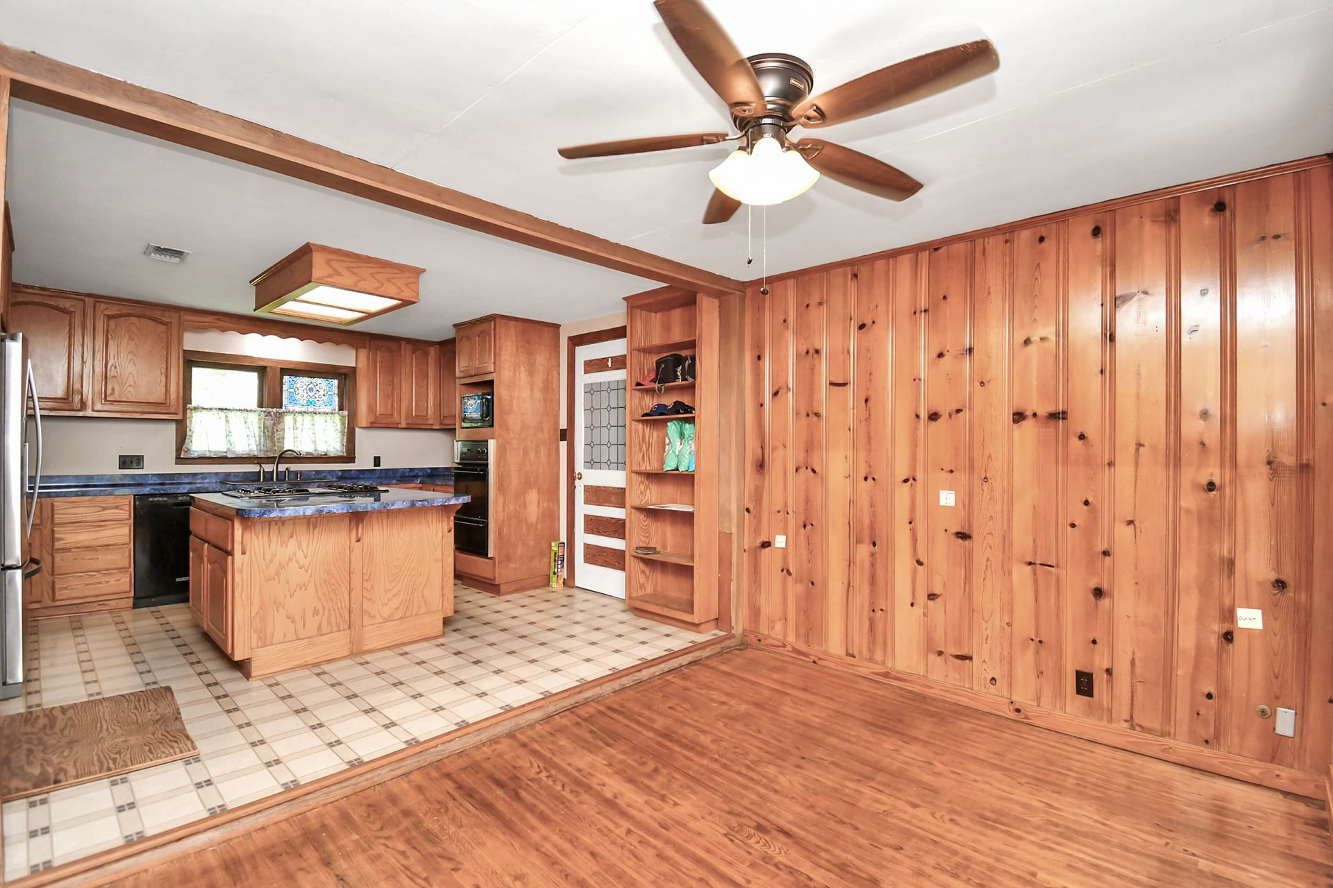 Kitchen with wood paneling, cabinets, and island. Flooring is tile and hardwood. Ceiling fan is present.