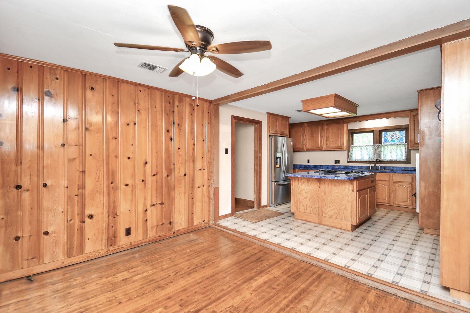 Wooden-paneled dining room opens to a kitchen with wood cabinets and blue countertops; a ceiling fan is present.