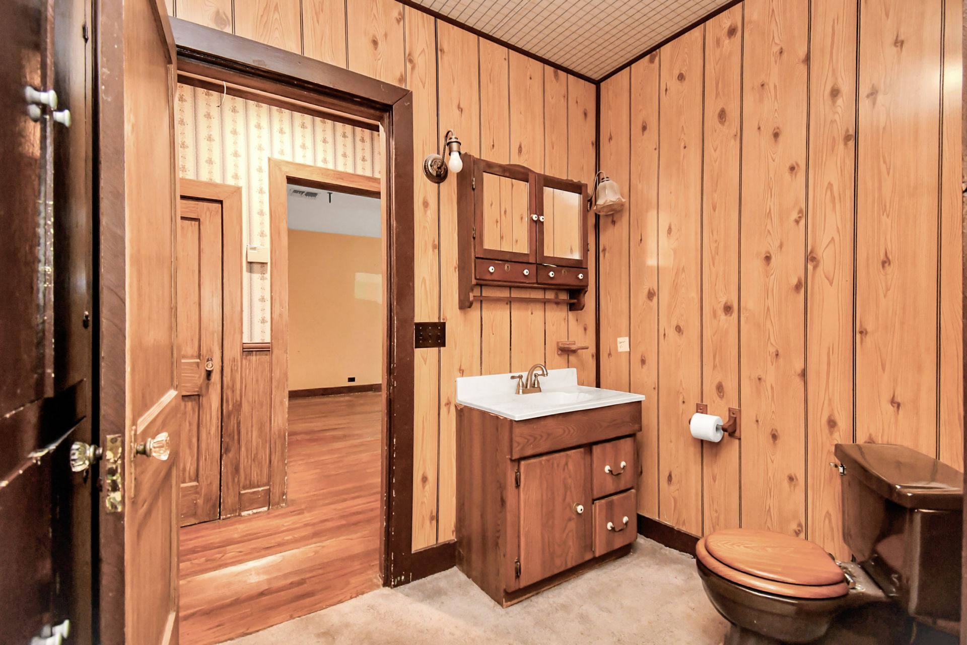 Bathroom with wood paneling, vanity, toilet, and an open doorway leading to a hallway and room.