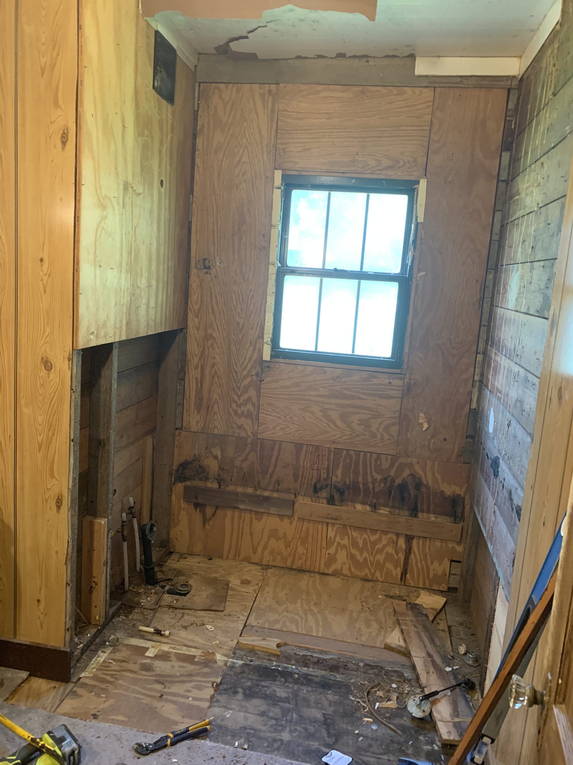 Bathroom renovation in progress: plywood walls, small window, exposed framing and plumbing.