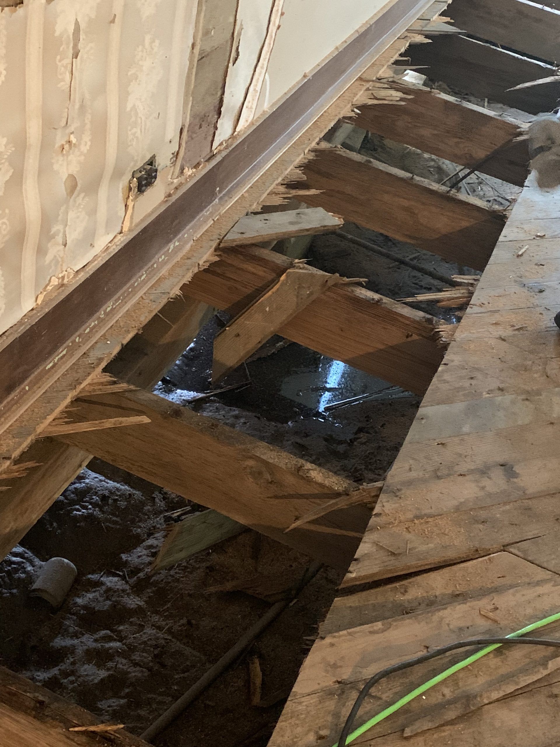 Damaged wooden floor with exposed beams, showing dark space below. Light reflects off water.