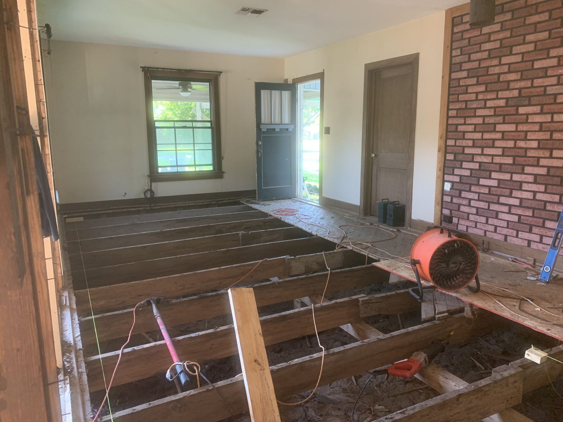 Interior room under renovation; exposed floor joists, brick accent wall, open doorway, and an electric fan.
