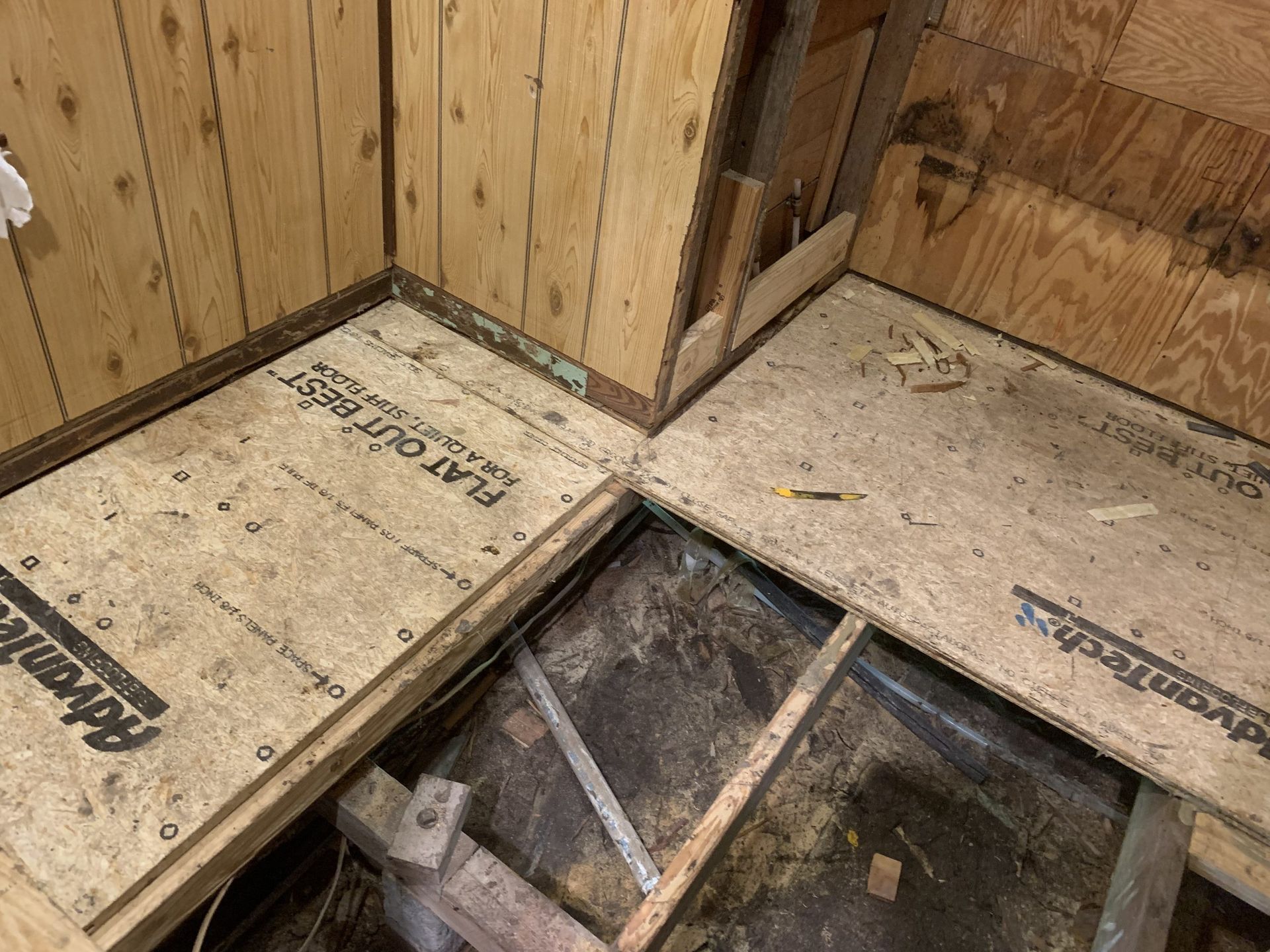 Interior of a room with plywood subfloor partially installed, revealing joists below. Brown paneling lines the walls.
