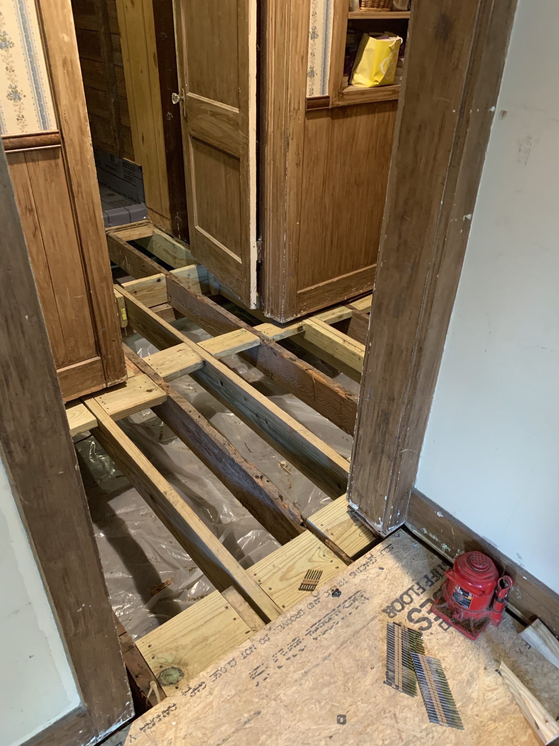 Hallway with exposed floor joists, plywood, plastic sheeting, and a red tool.