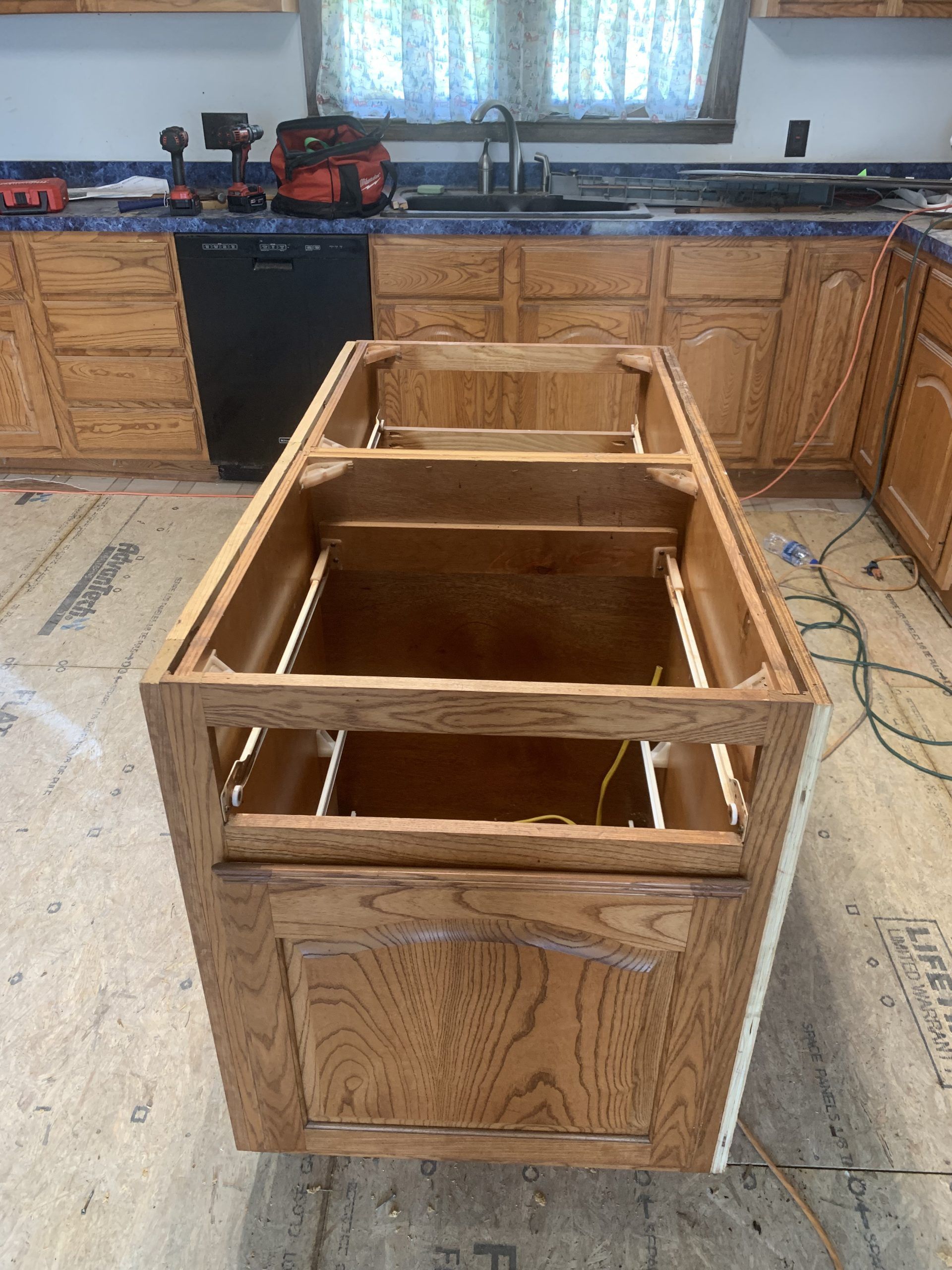 Oak kitchen island cabinet with missing countertop, set in a room with other cabinets and countertop.