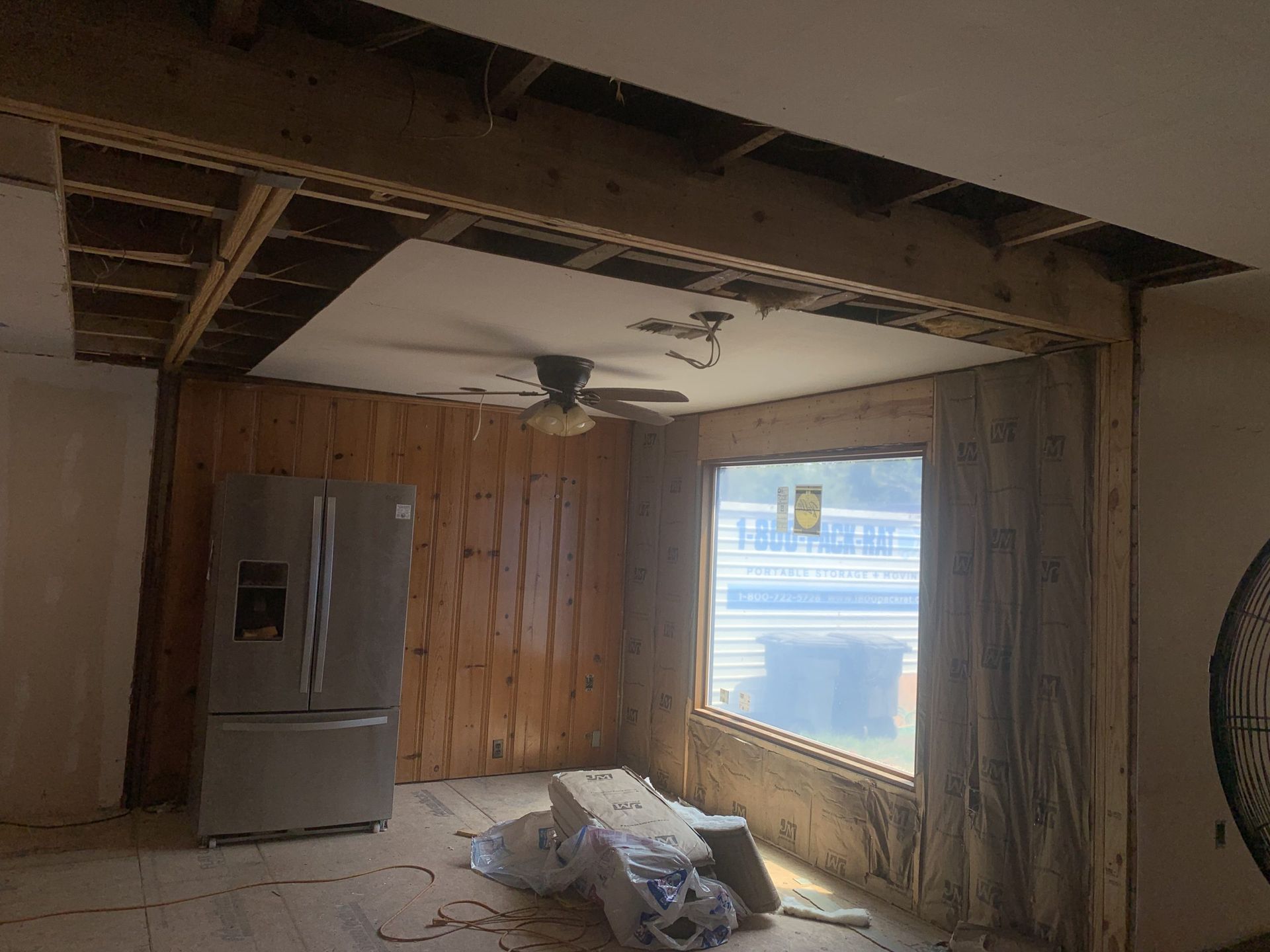 Room under renovation with wood paneling, exposed ceiling beams, refrigerator, and window.