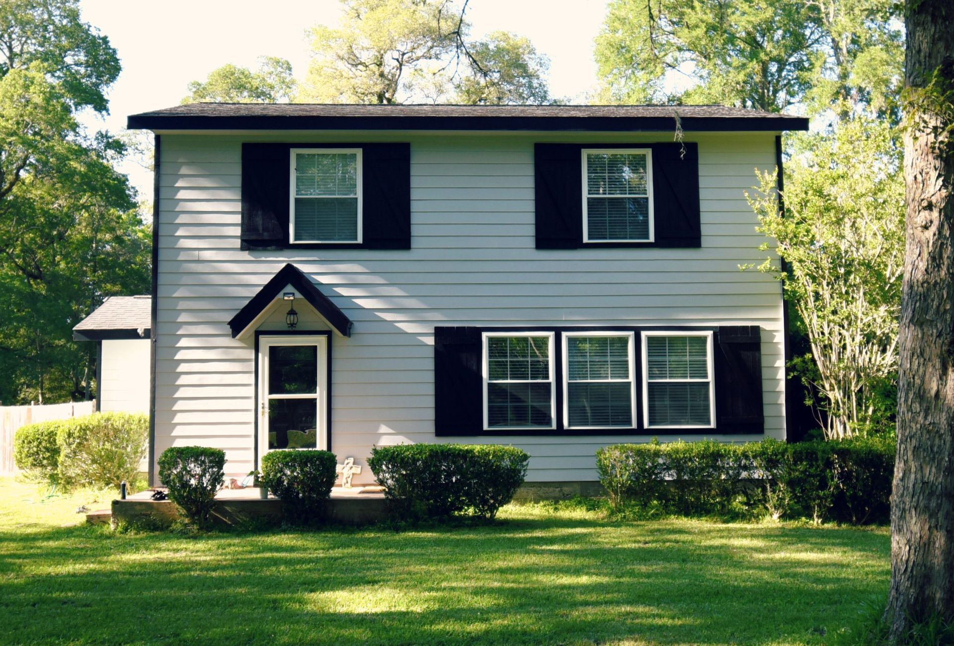 Two-story light gray house with black shutters and windows on a grassy lawn, surrounded by trees.