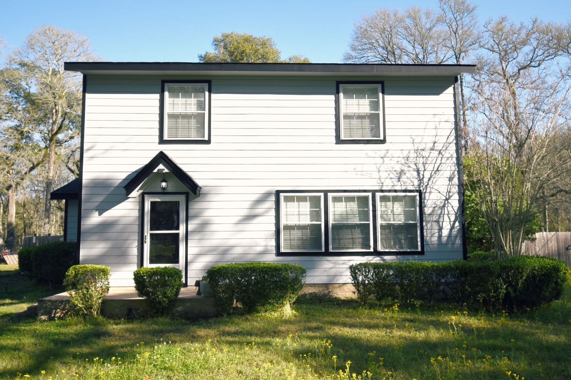 Two-story light blue house with black trim and a small porch, surrounded by bushes and trees.
