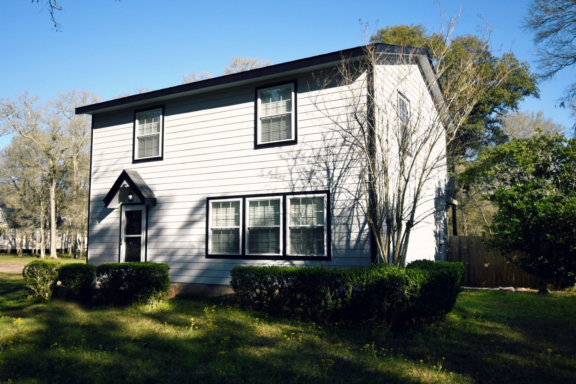 Two-story house with light siding, black trim, and manicured bushes in front.