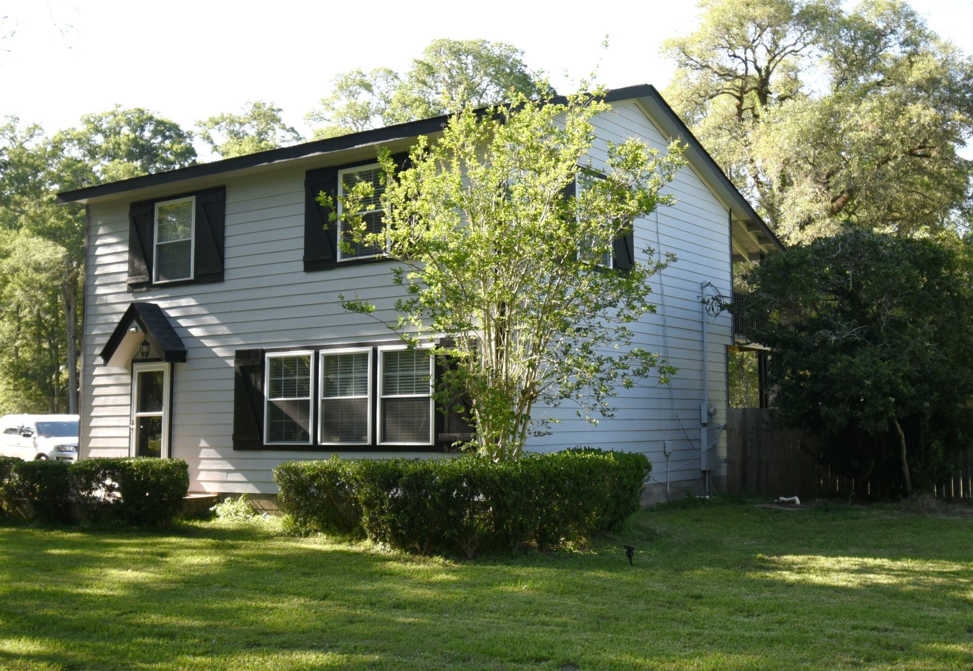 Two-story light gray house with black shutters and trim, fronted by a green lawn and bushes.