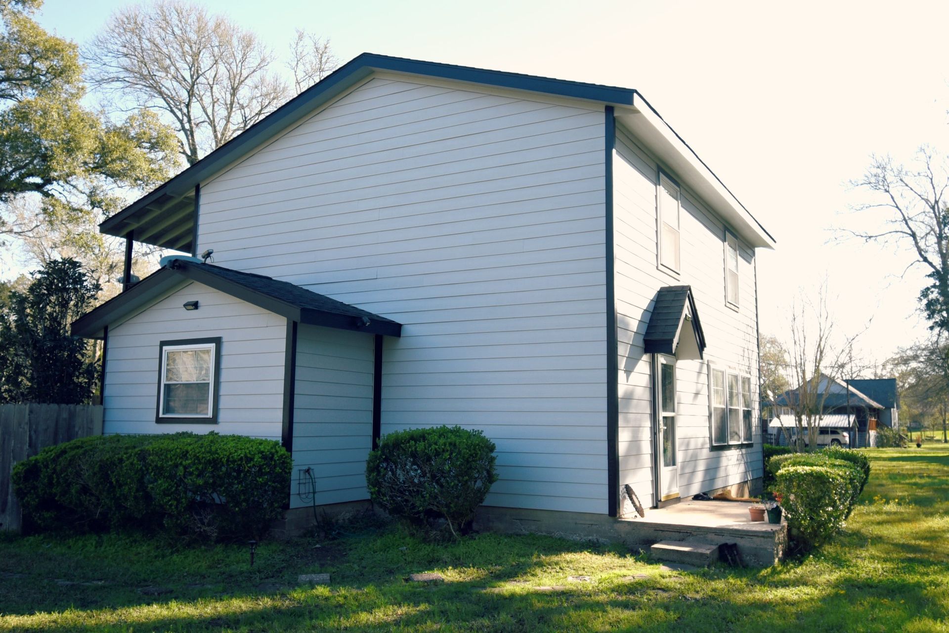 Two-story light blue house with a small covered porch and a small attached shed. Green bushes and grass surround the house.