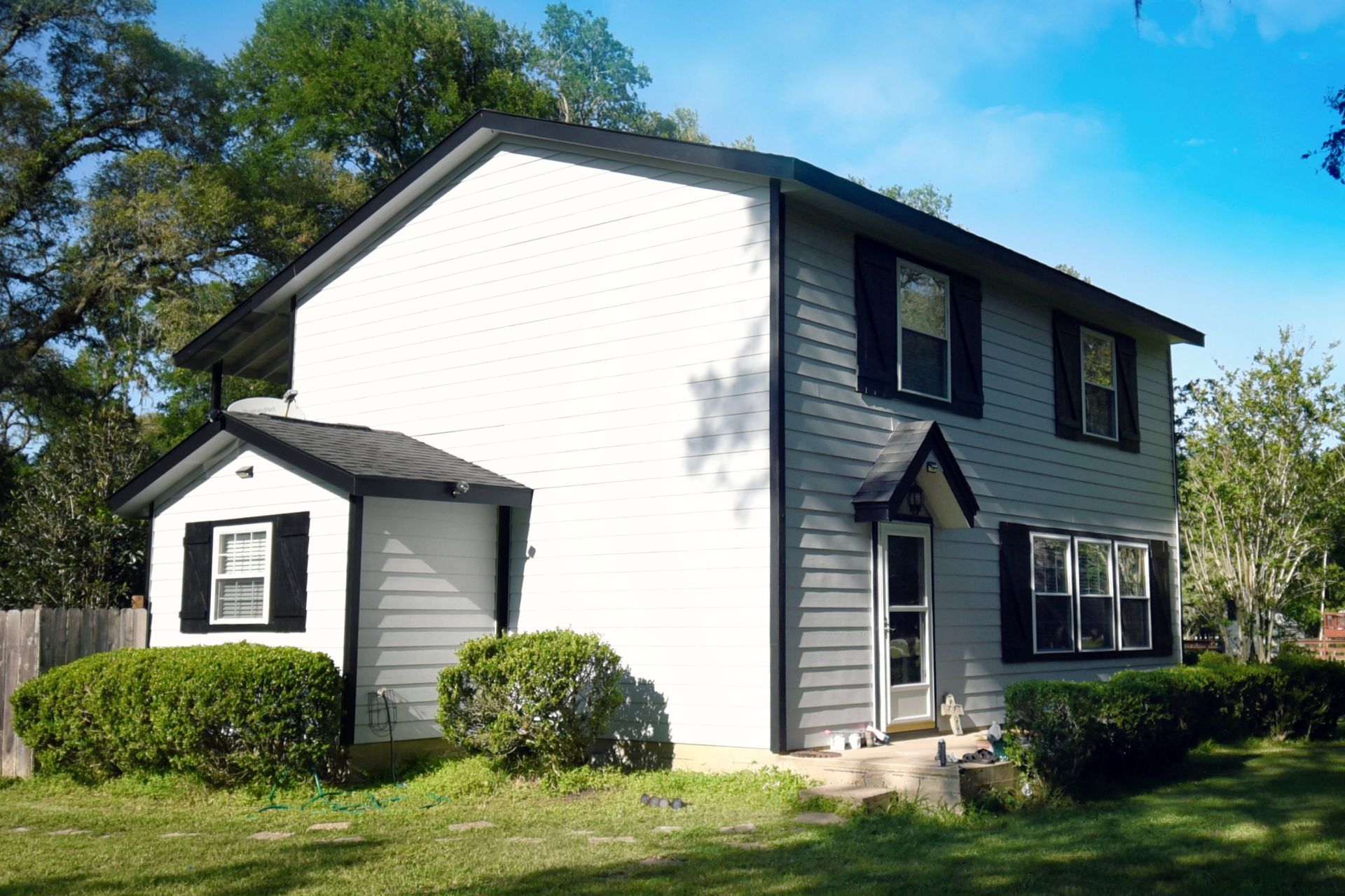 Two-story gray house with black shutters and roof, green bushes, and a sunny sky.