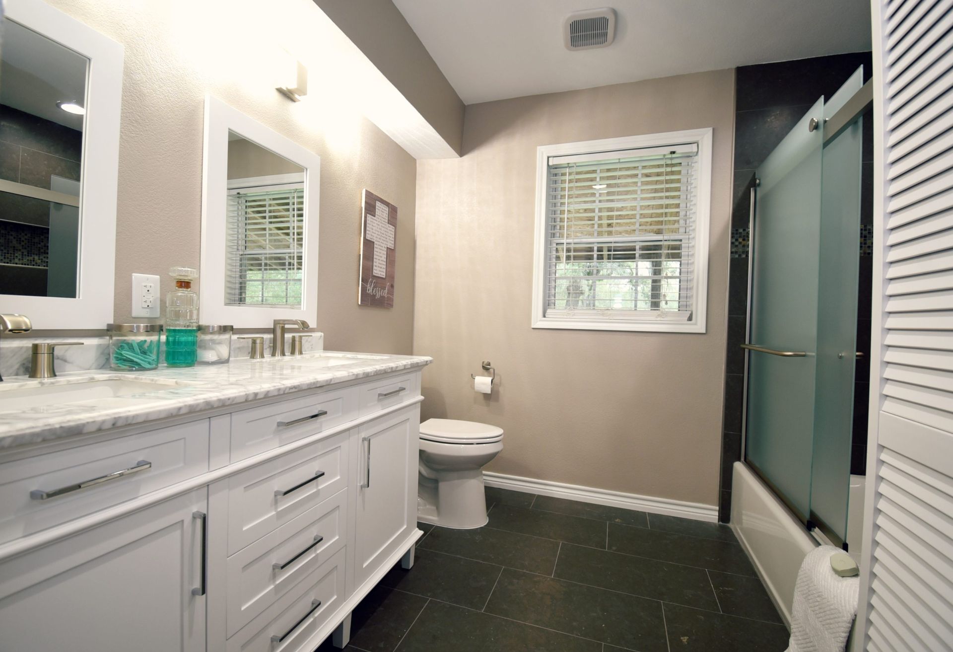 Bathroom with white double vanity, toilet, shower, and window with blinds. Beige walls, dark tiled floor.