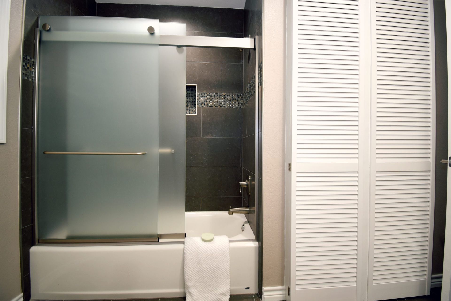 Bathroom with frosted glass shower door over a white tub, next to white louvered door.