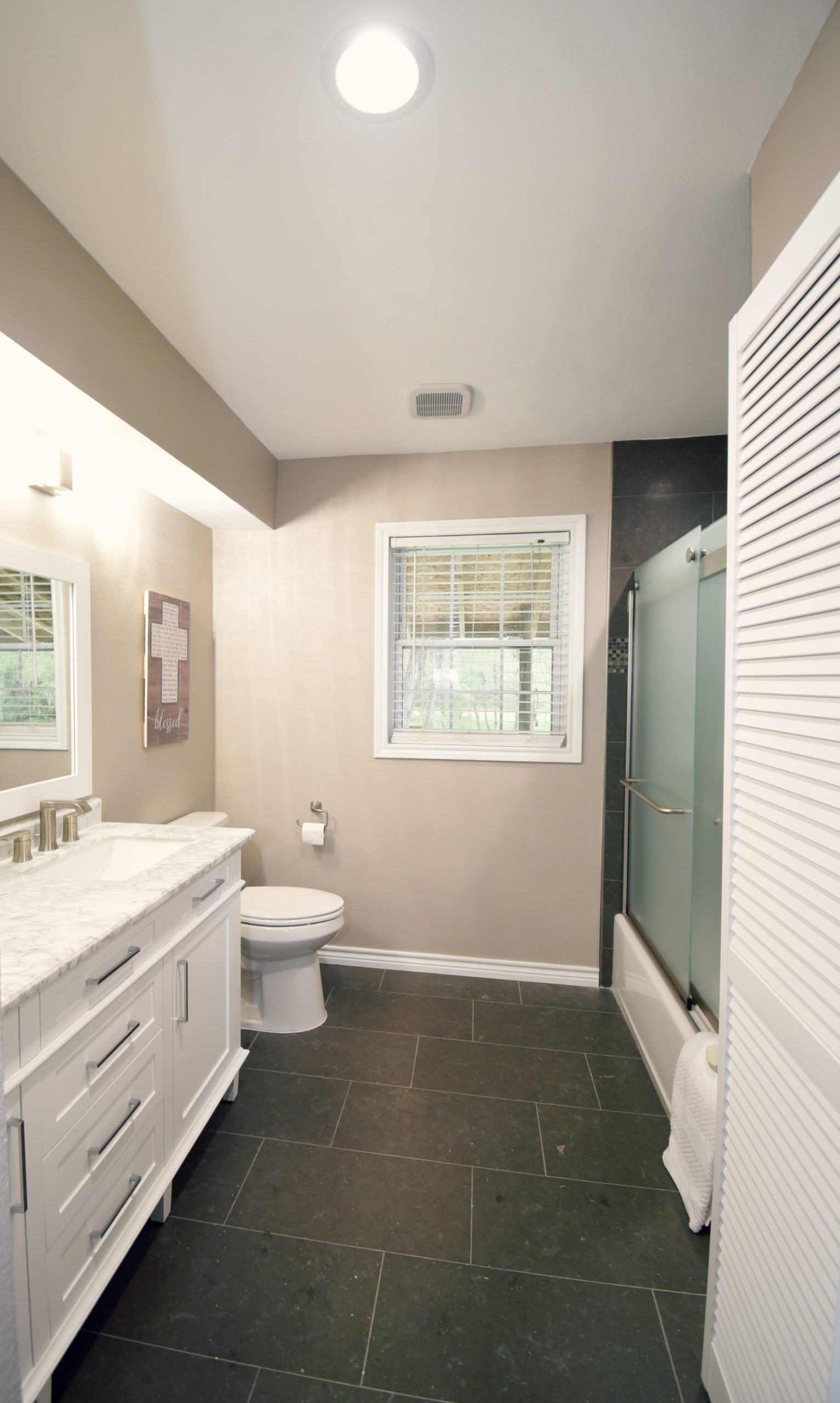 Bathroom with a white vanity, toilet, shower, and shuttered door. Dark floor, beige walls, window.