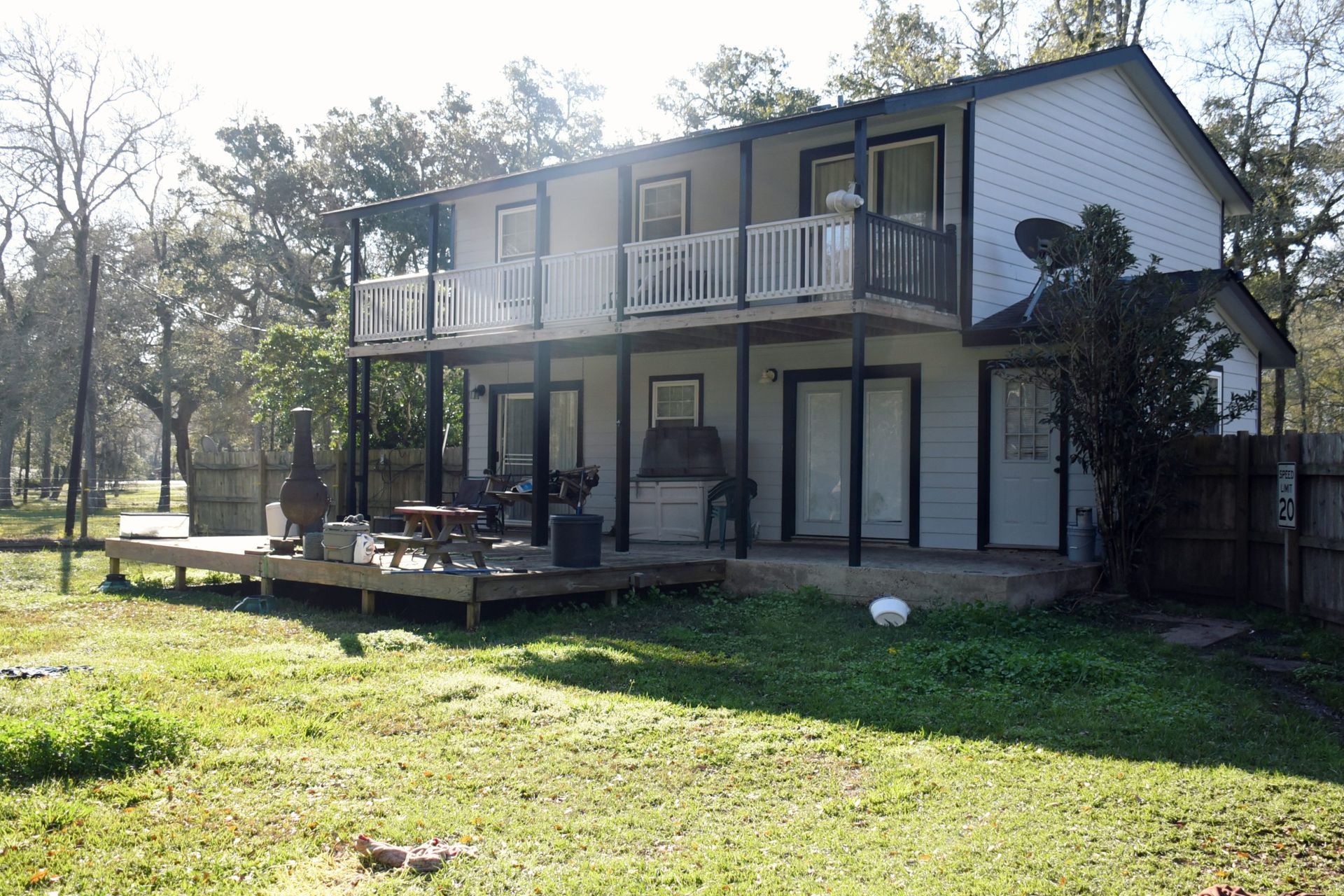 Two-story house with balconies, wooden deck, and yard. Light-colored walls and dark trim. Trees in background.