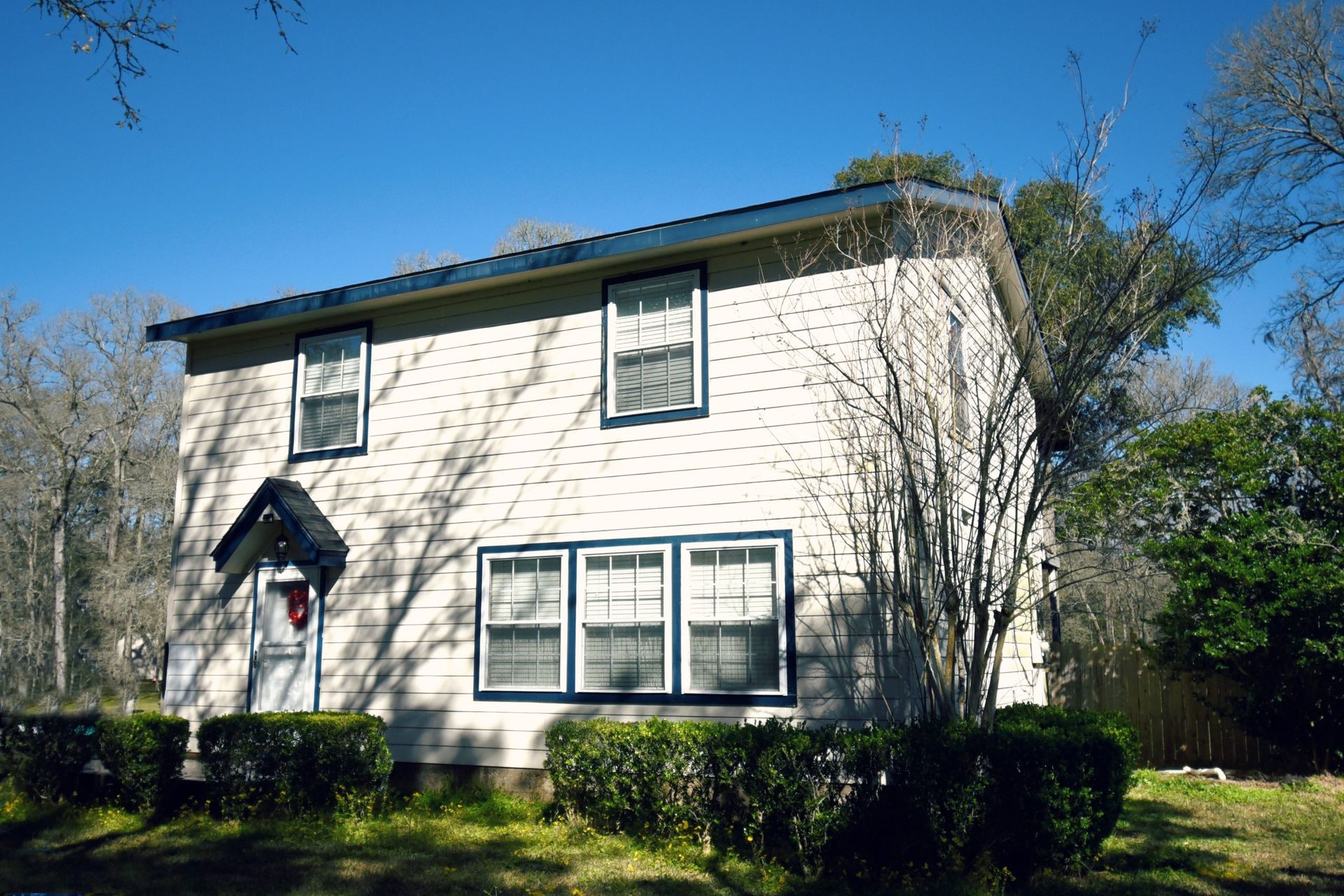 Two-story beige house with blue trim and windows, set in a yard with greenery and trees on a sunny day.