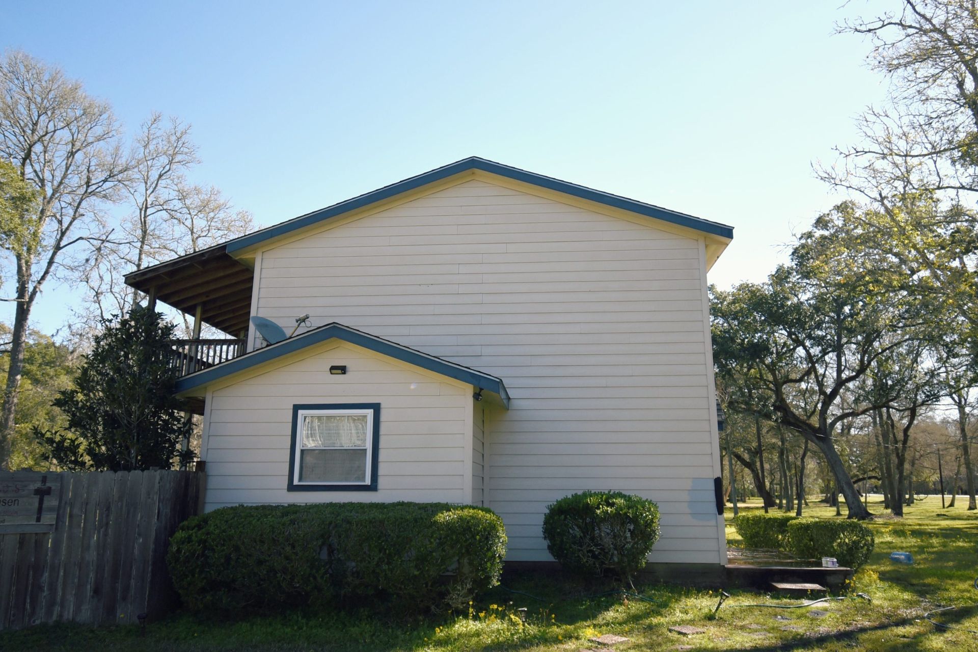 Two-story house with light-colored siding, a green-trimmed roof, and small shed.  Green bushes in front. Sunny day.