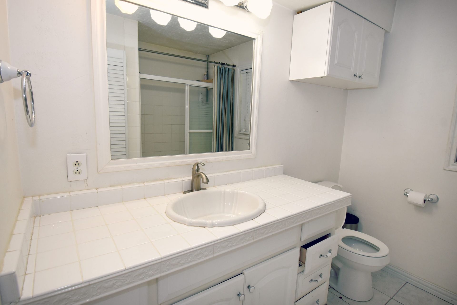 Bathroom with white vanity, sink, and toilet; overhead cabinet. Mirror reflects shower stall.