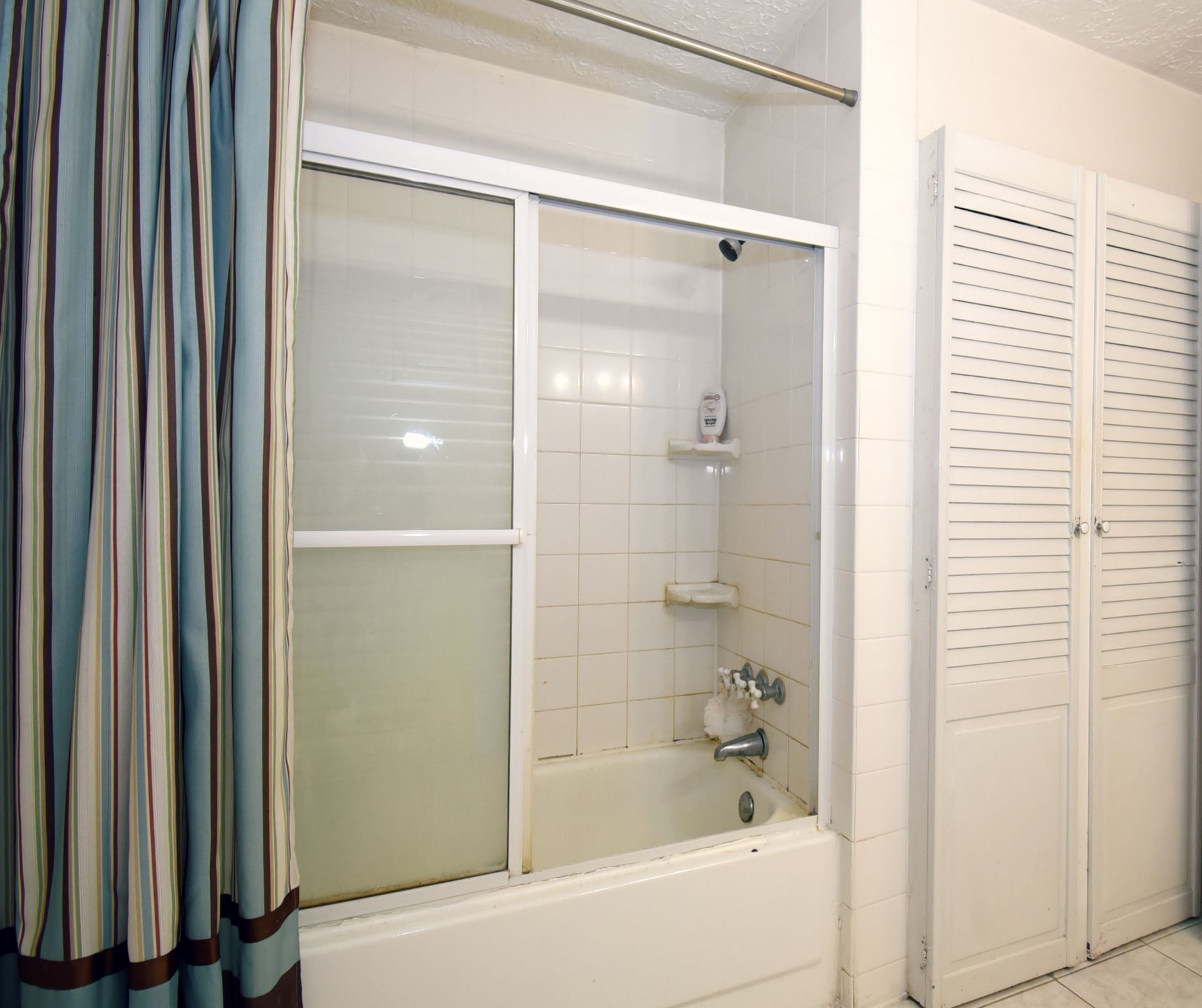 Bathroom with shower/tub, white tile, sliding glass door, blue and white curtain, and white closet.